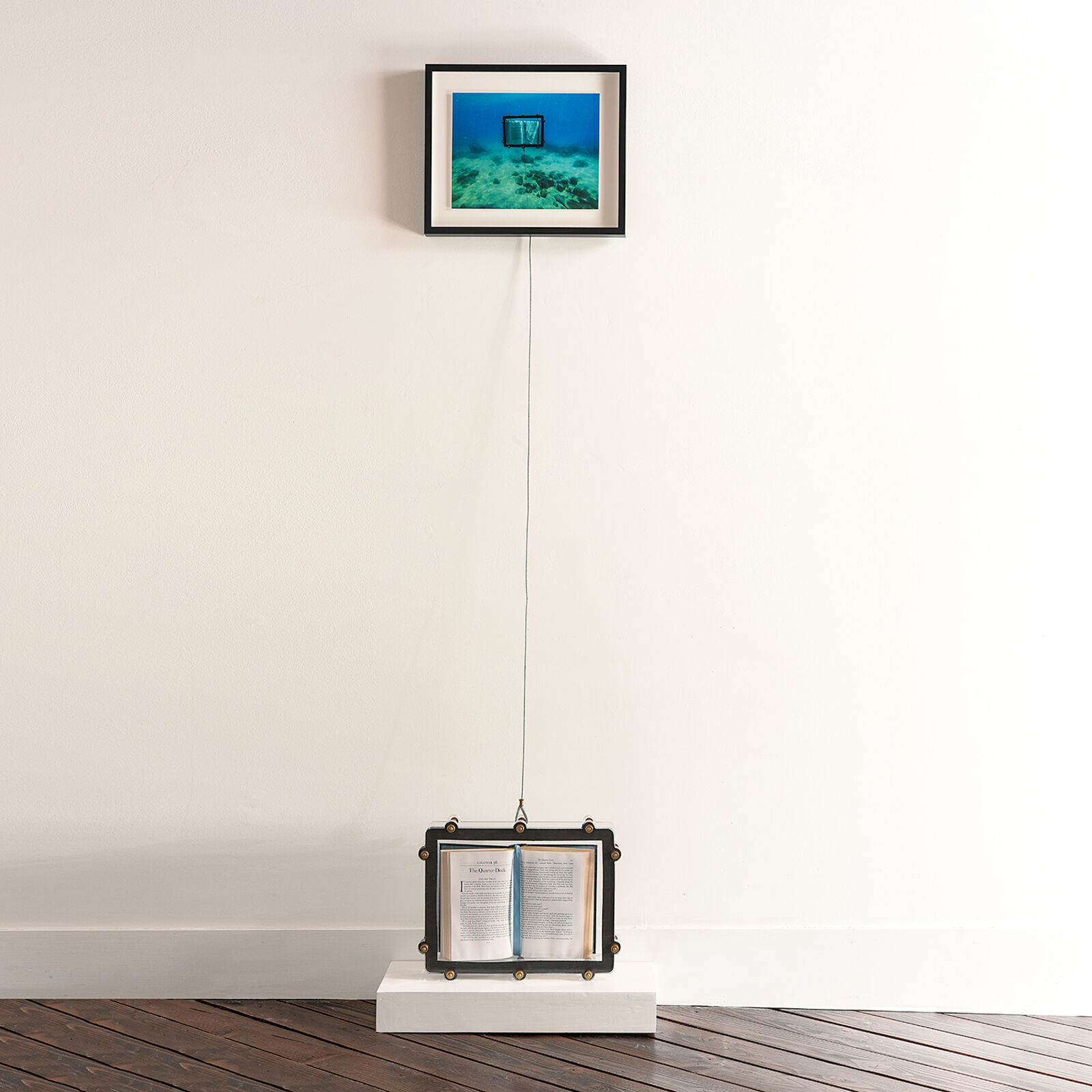A surreal installation featuring an open book suspended by a wire from a framed underwater photograph.