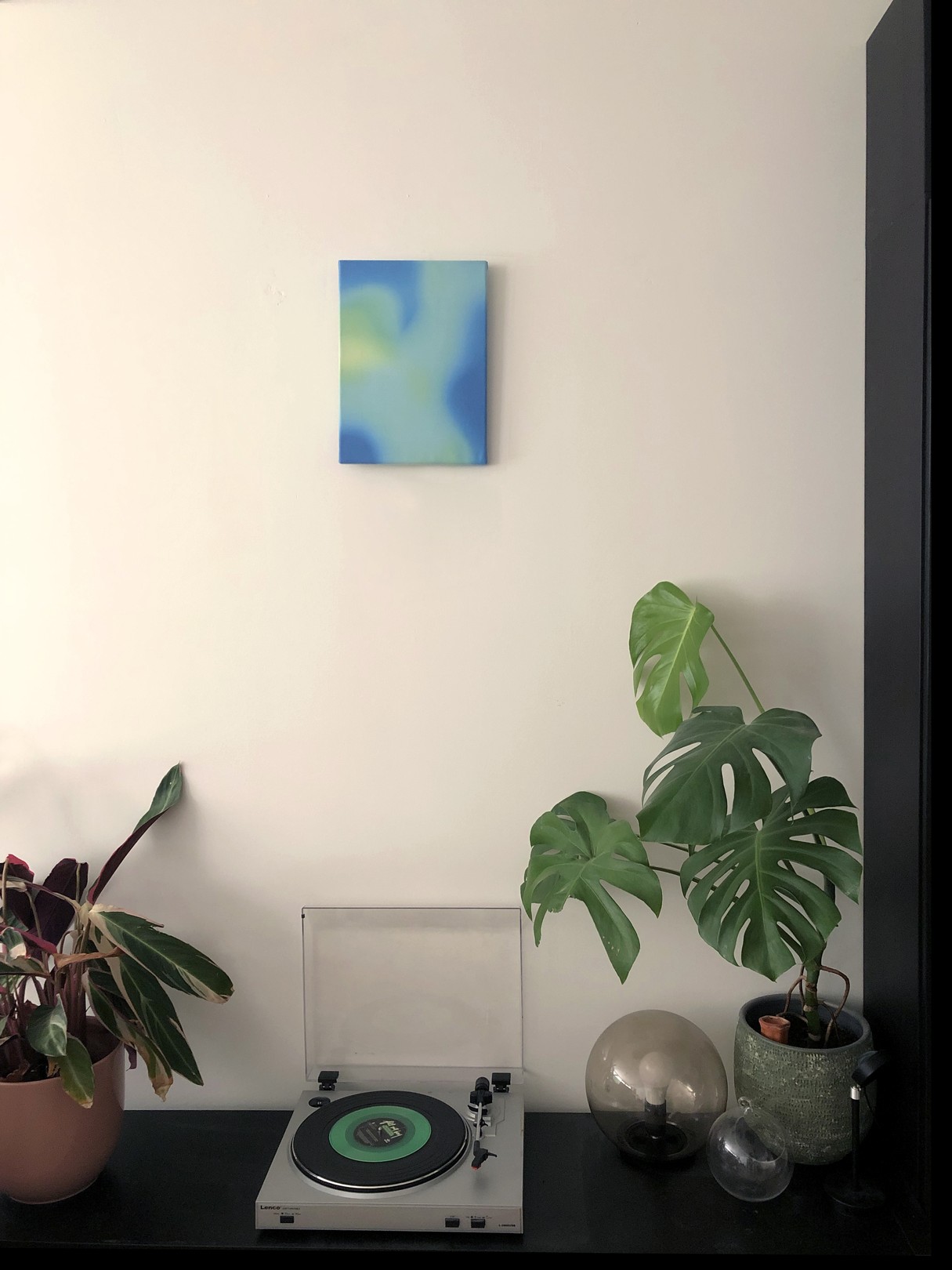 A minimalist abstract painting featuring soft blue and green gradients on a white wall.