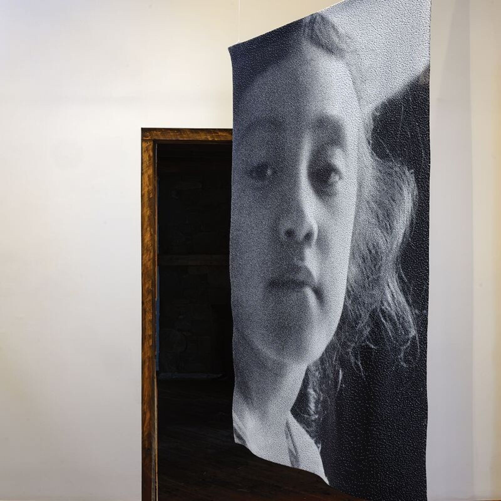 A black-and-white portrait of a person with wavy hair, framed by a wooden doorway.