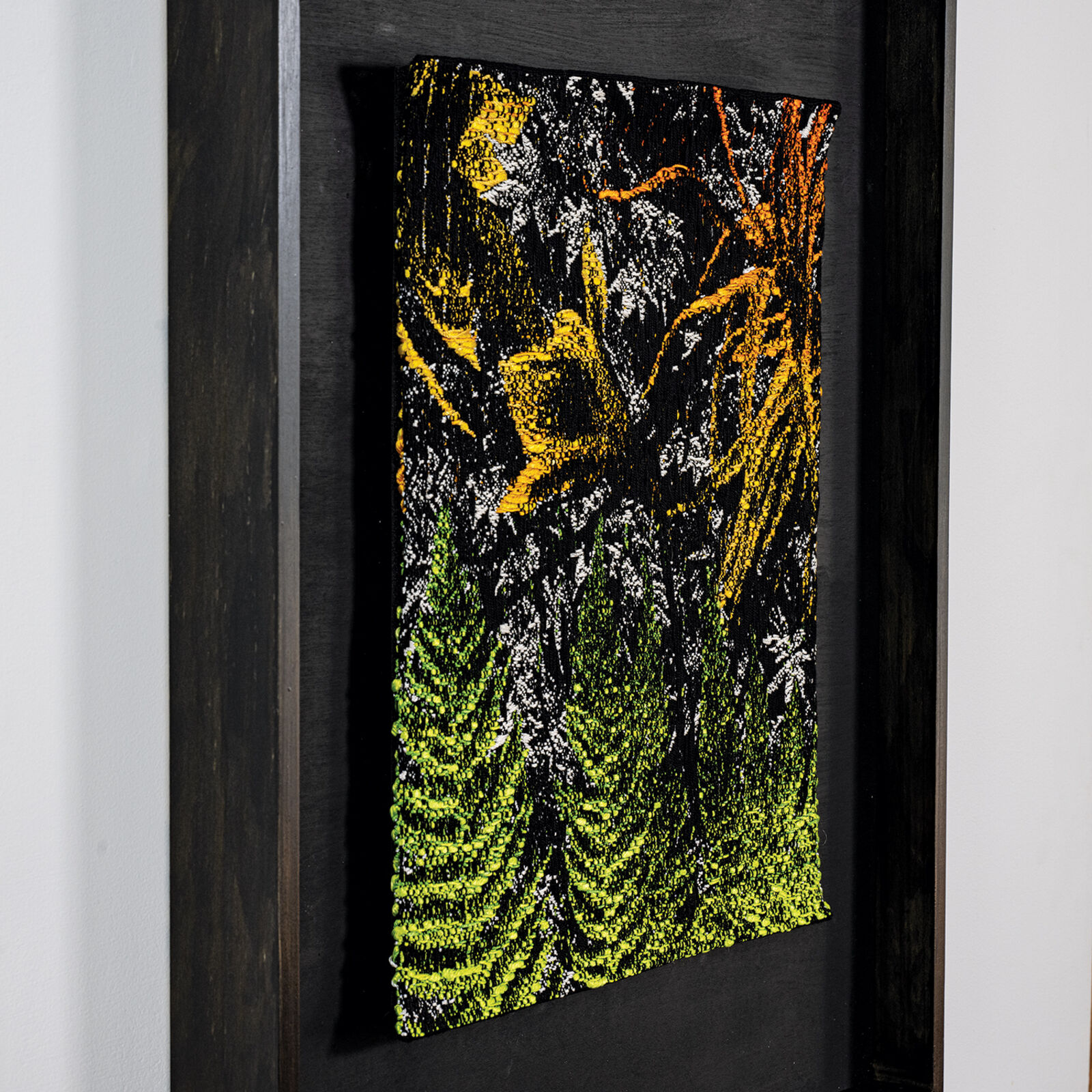 A framed textile artwork featuring a dense, textured forest scene with vibrant green ferns and yellow foliage against a dark background.