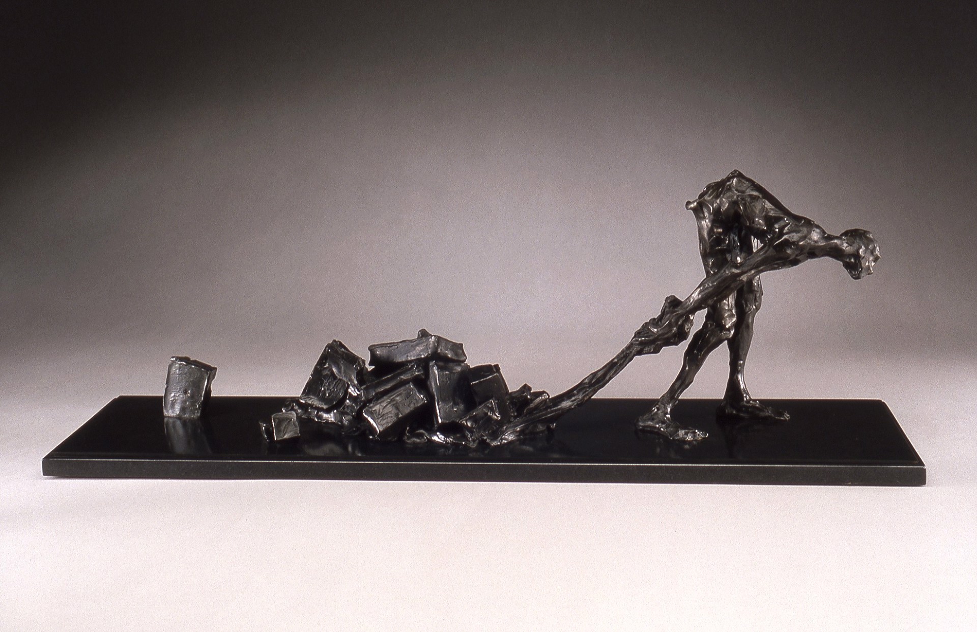 A dark metal sculpture of a bent figure dragging a heavy load of blocks across a flat base.