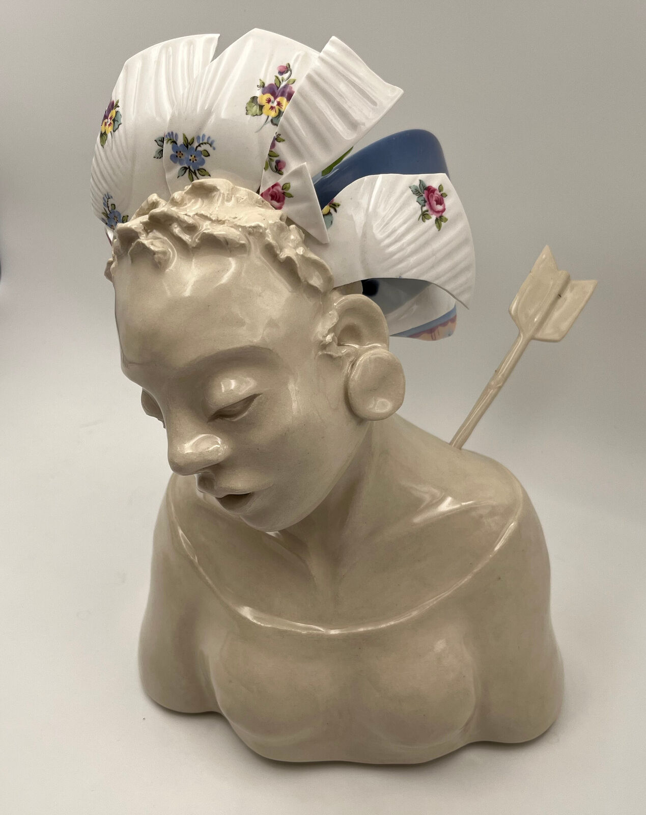 A ceramic bust of a figure pierced by an arrow, wearing a floral bonnet and resting against a plain background.