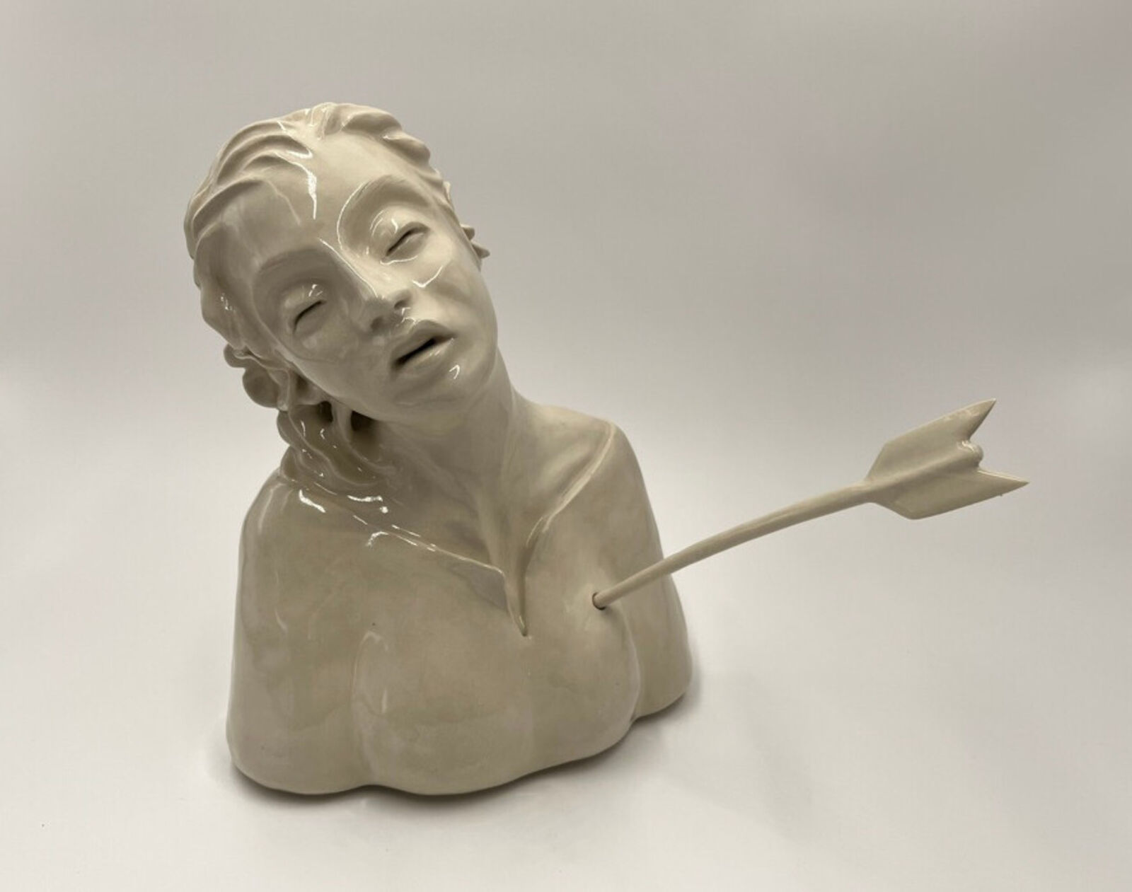A glossy ceramic bust of a figure pierced by an arrow, rendered in monochromatic tones against a neutral background.