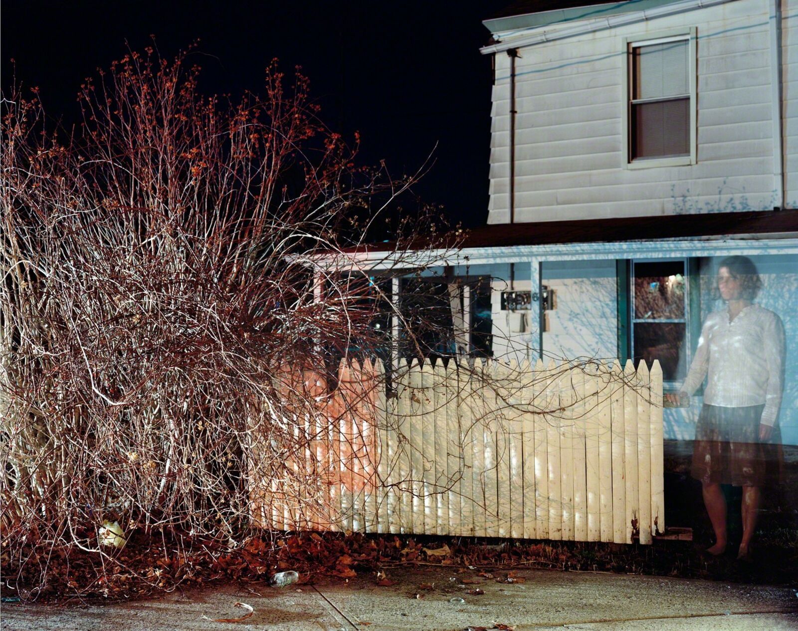 A ghostly figure stands in a window of a weathered house, framed by overgrown branches and a picket fence.
