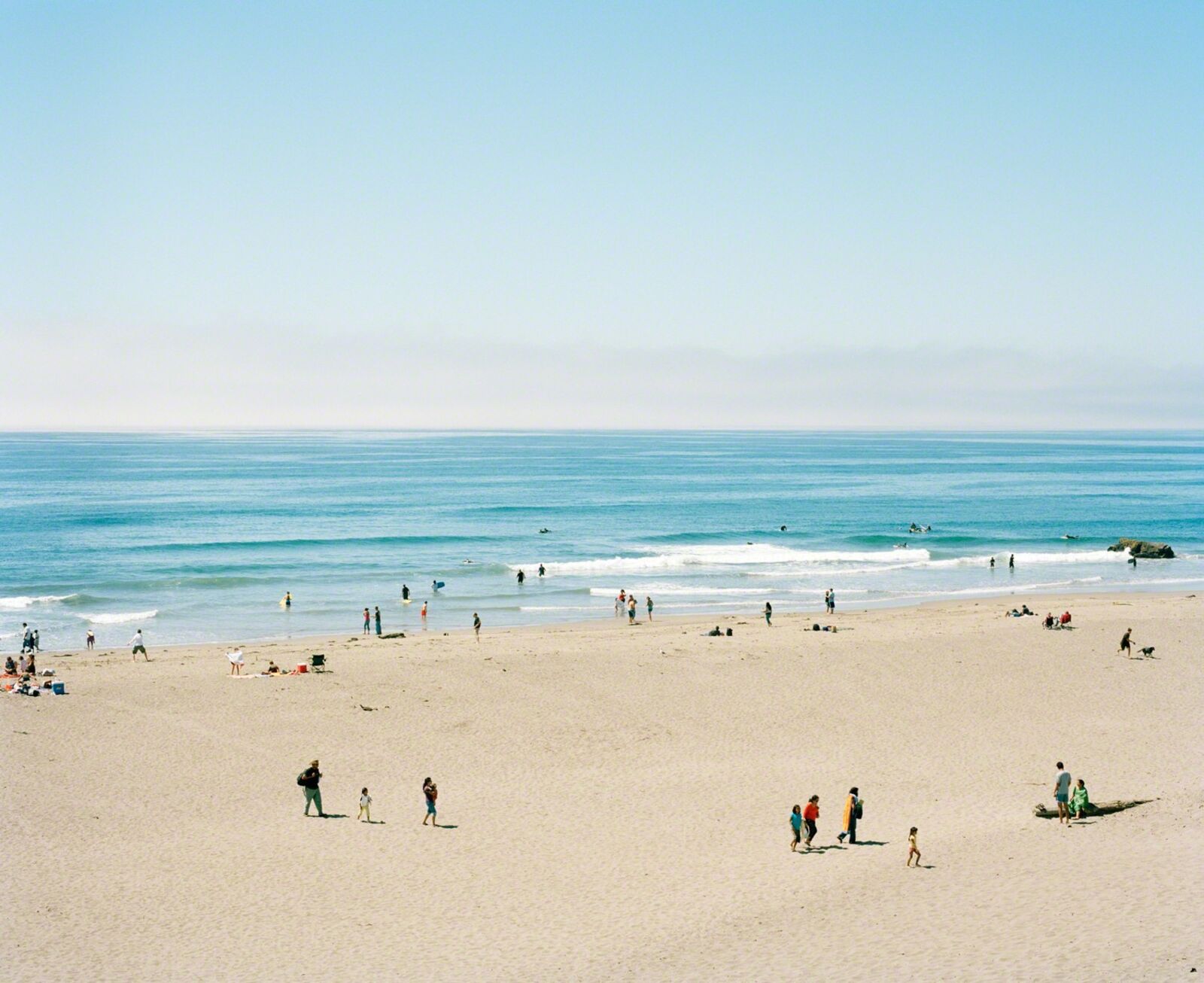 A wide-angle view of a sunlit beach with calm blue water, sandy shore, and scattered figures enjoying the day.