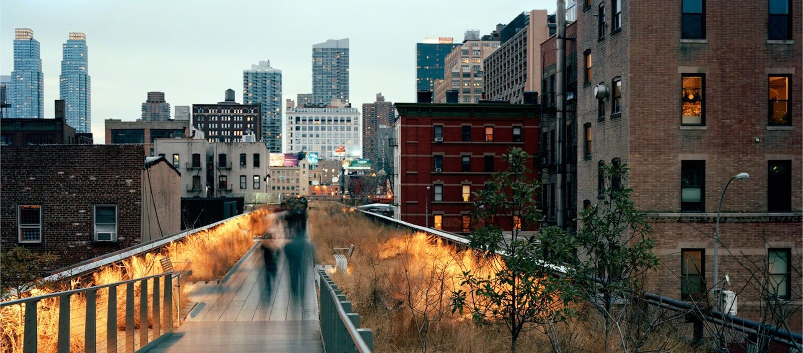 A long-exposure photograph of a city rooftop garden with blurred figures walking on a wooden path.