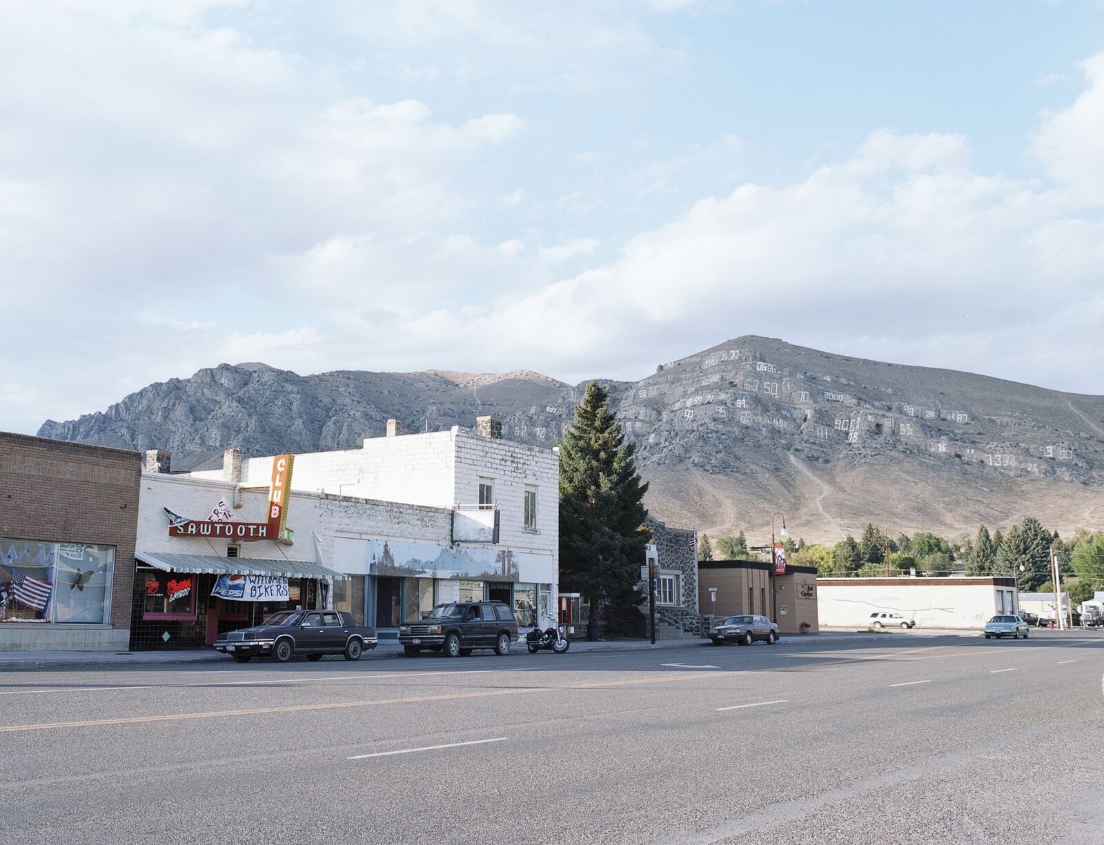 A quiet roadside scene featuring a row of storefronts and parked vehicles against a backdrop of mountains with large numbers inscribed on the slopes.