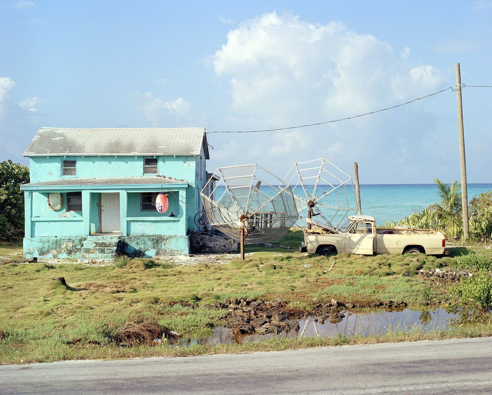A weathered turquoise house stands beside a large satellite dish and an abandoned truck near the ocean.