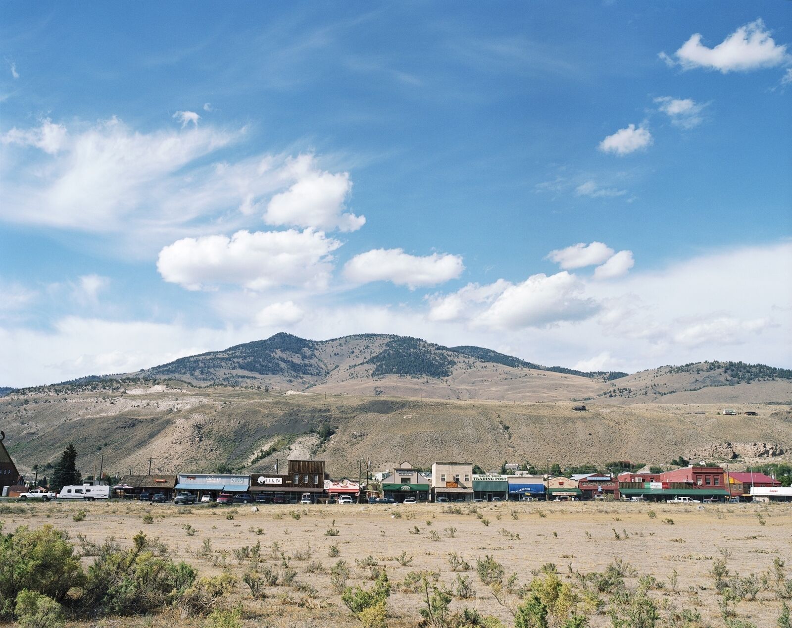 A wide-angle landscape photograph of a small town nestled at the foot of a large, tree-dotted mountain under a vast blue sky.