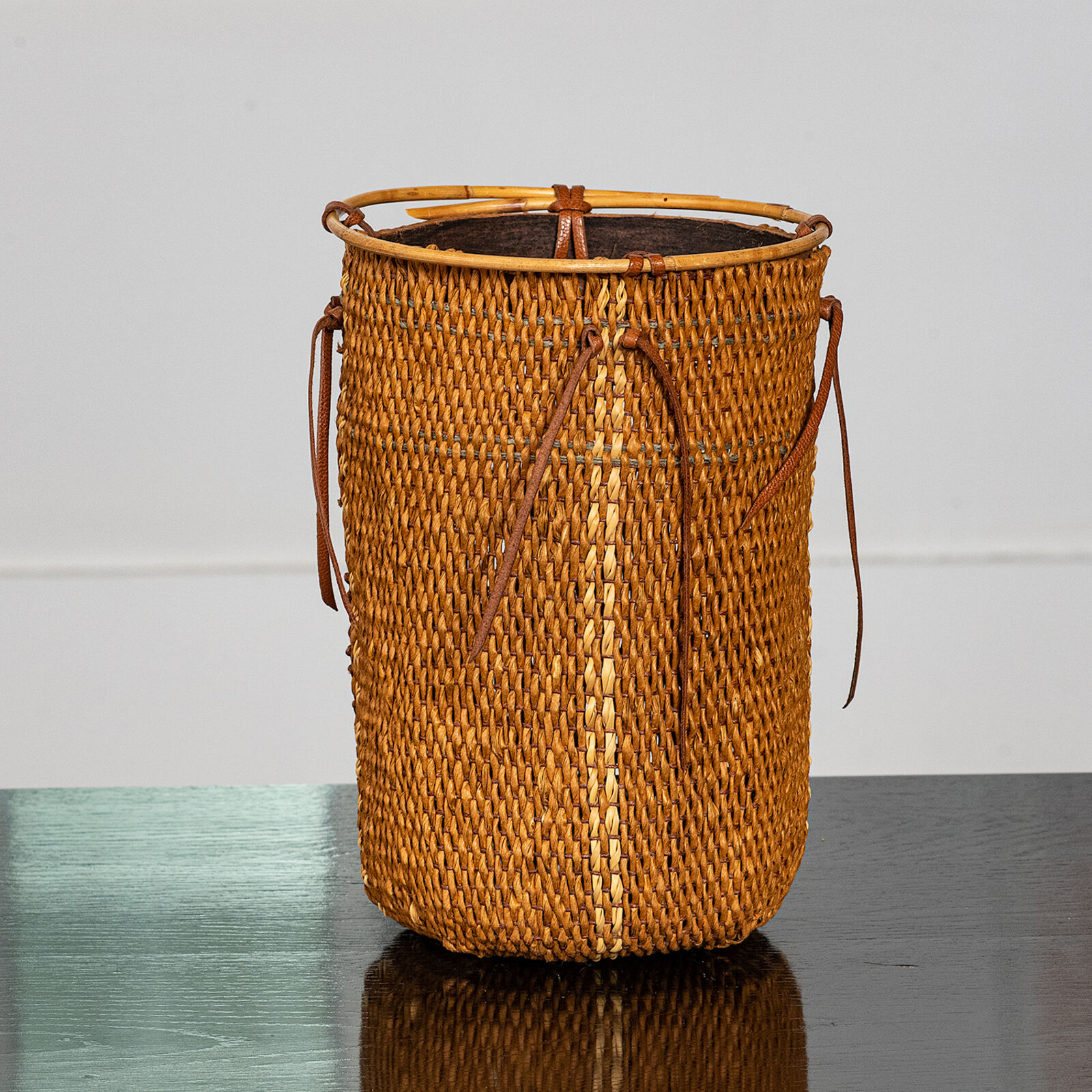 A woven basket with leather straps and a bamboo rim sits on a reflective surface.