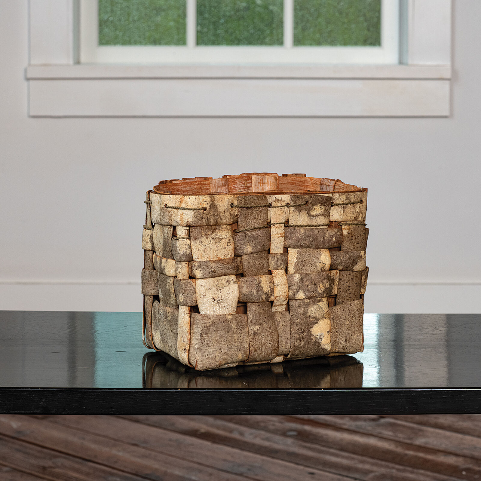 A woven wooden basket sits on a glossy black surface near a window.