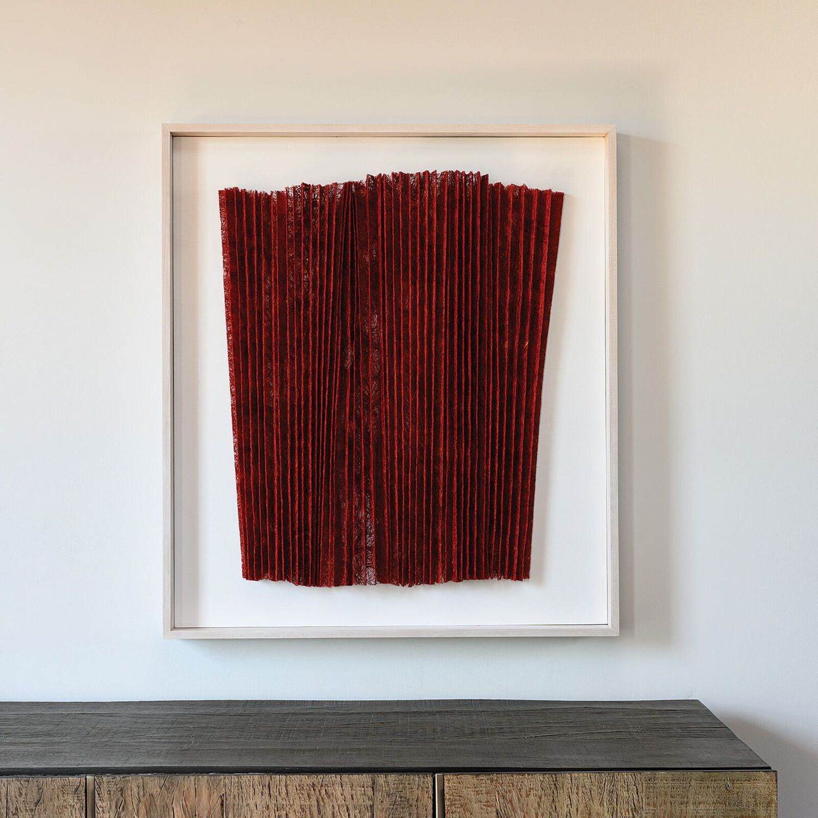 A minimalist textile artwork featuring deep red pleated fabric in a light frame.