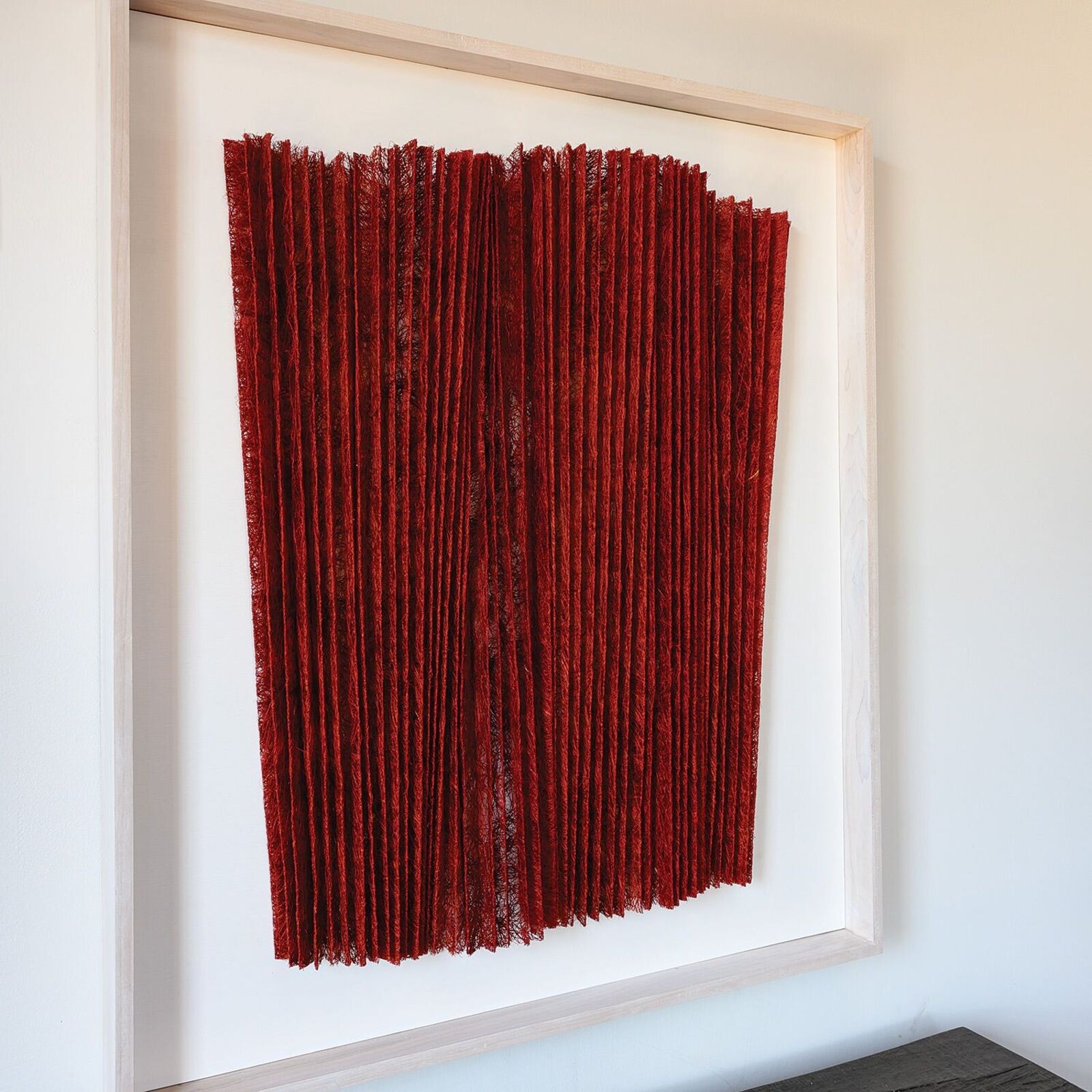 A minimalist textile artwork featuring deep red pleated fabric in a light frame.