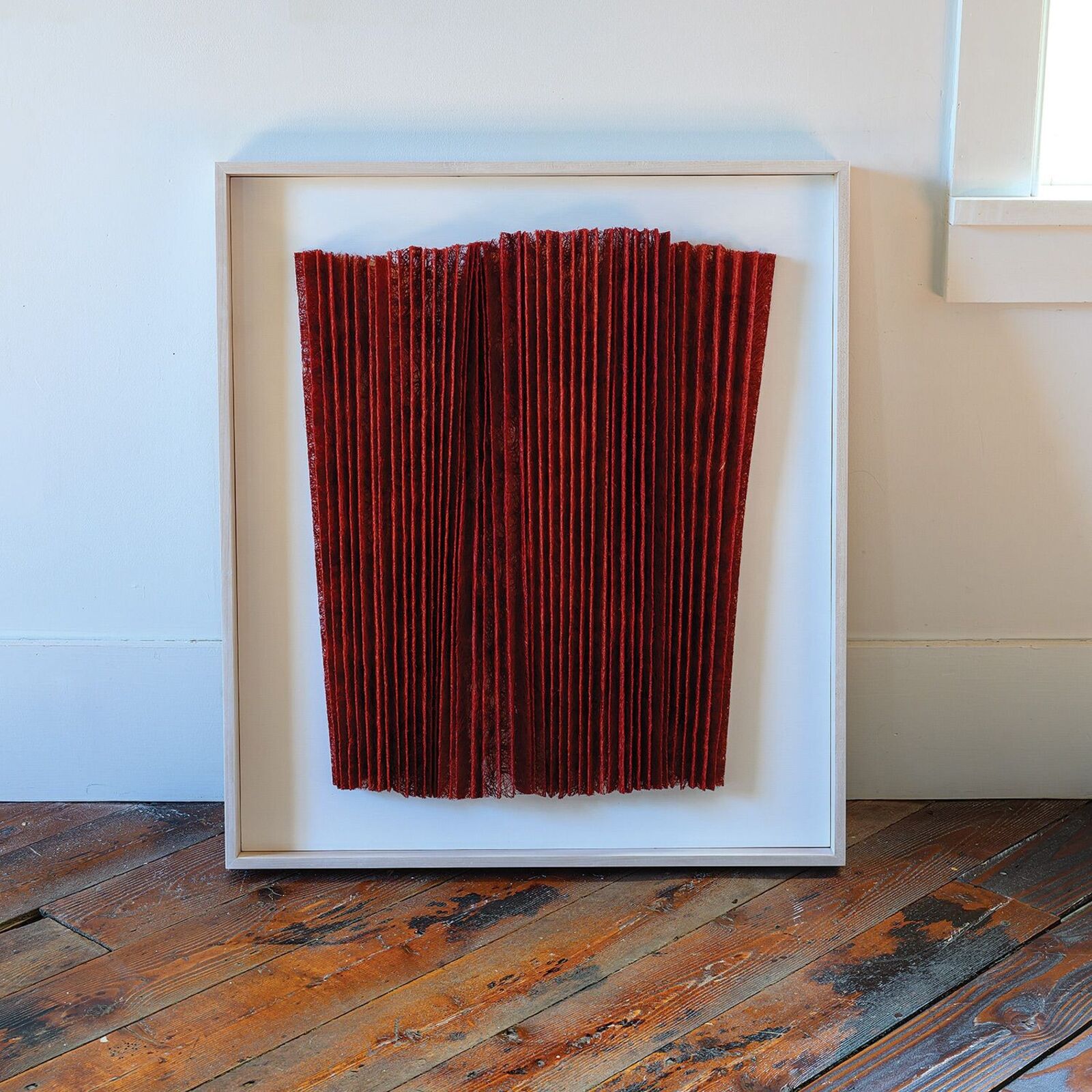 A minimalist textile artwork featuring deep red pleated fabric in a light frame.