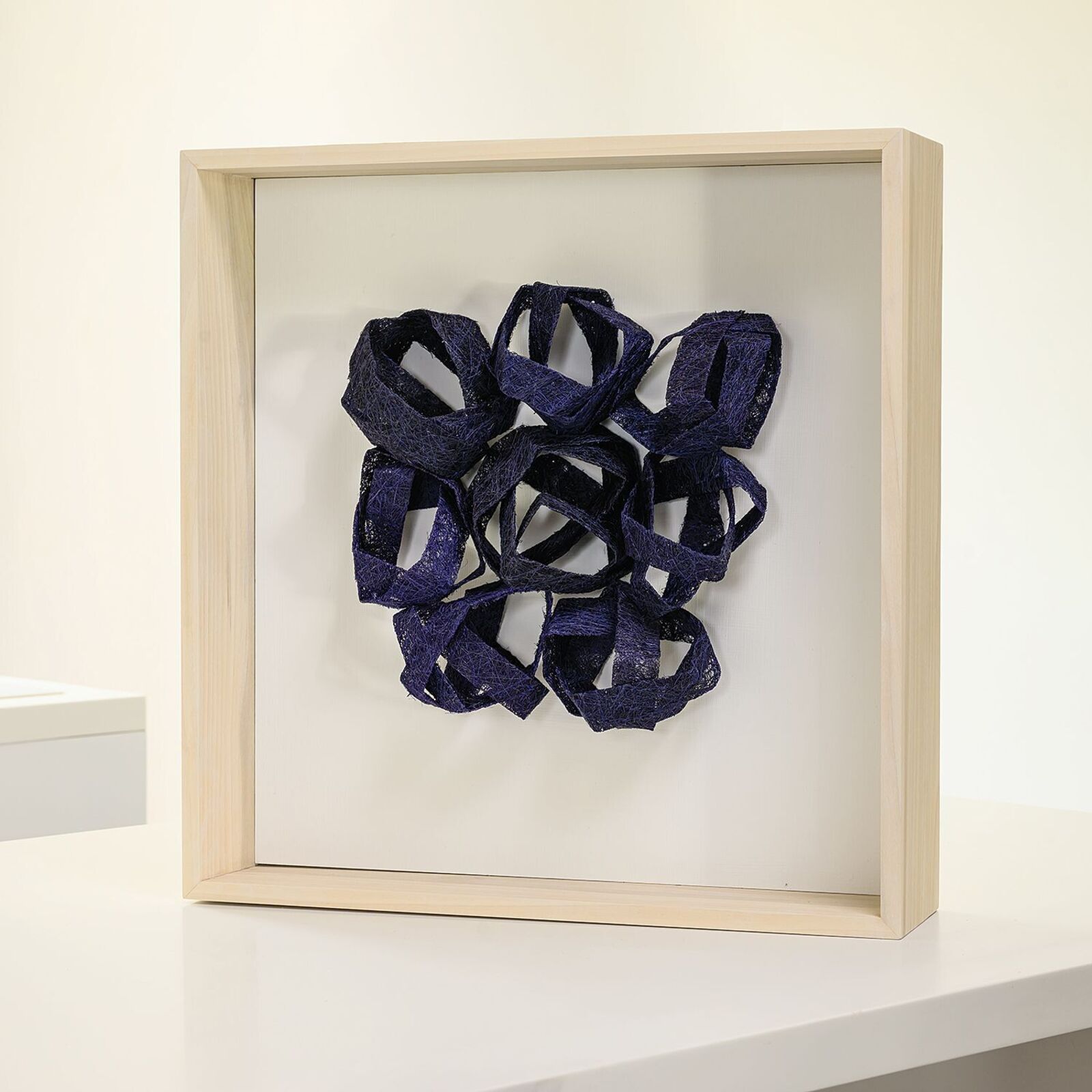 A framed textile sculpture featuring interwoven dark blue ribbons.