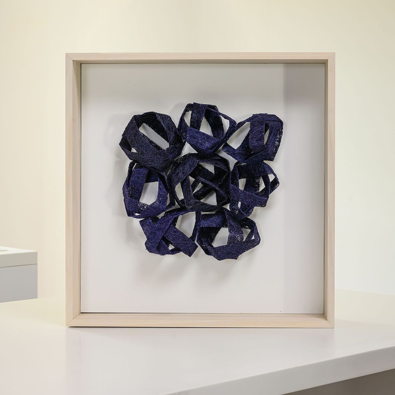 A framed textile sculpture featuring interwoven dark blue ribbons.