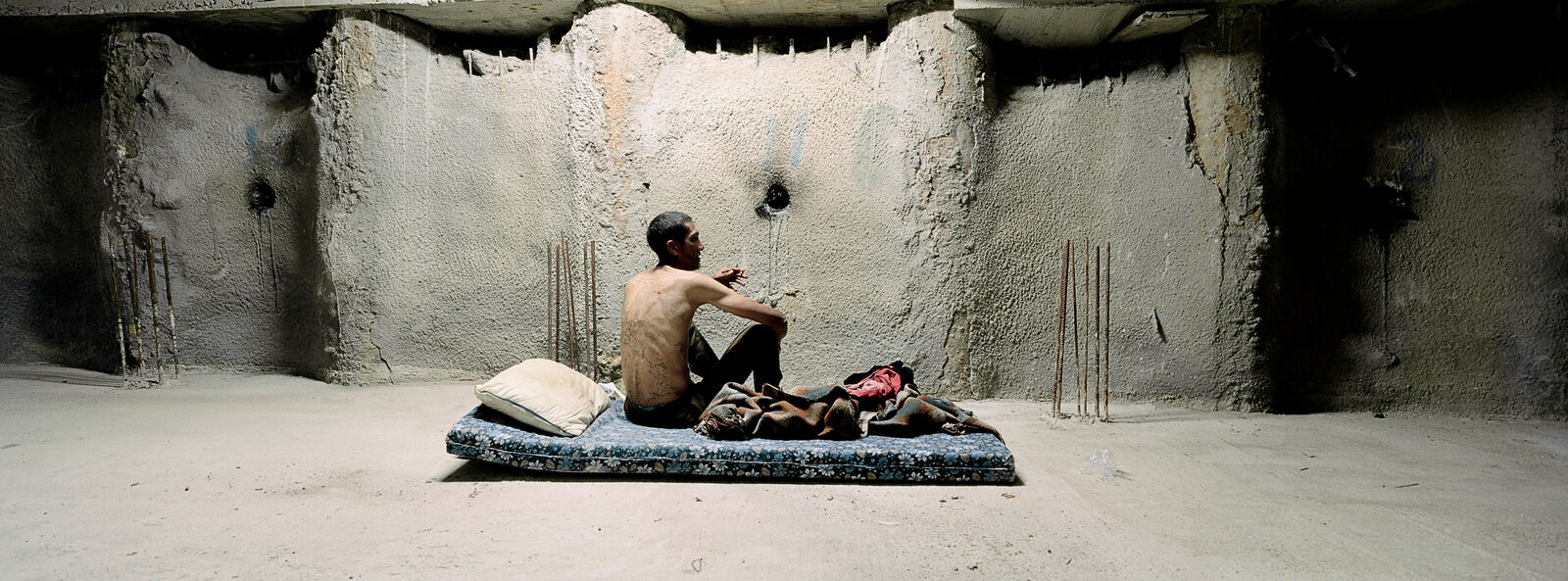 A man sitting on a bed in a room with concrete walls.