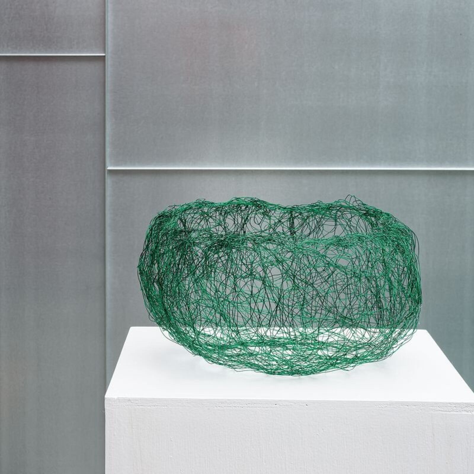 A green wire sculpture resembling a nest sits in a white display case.