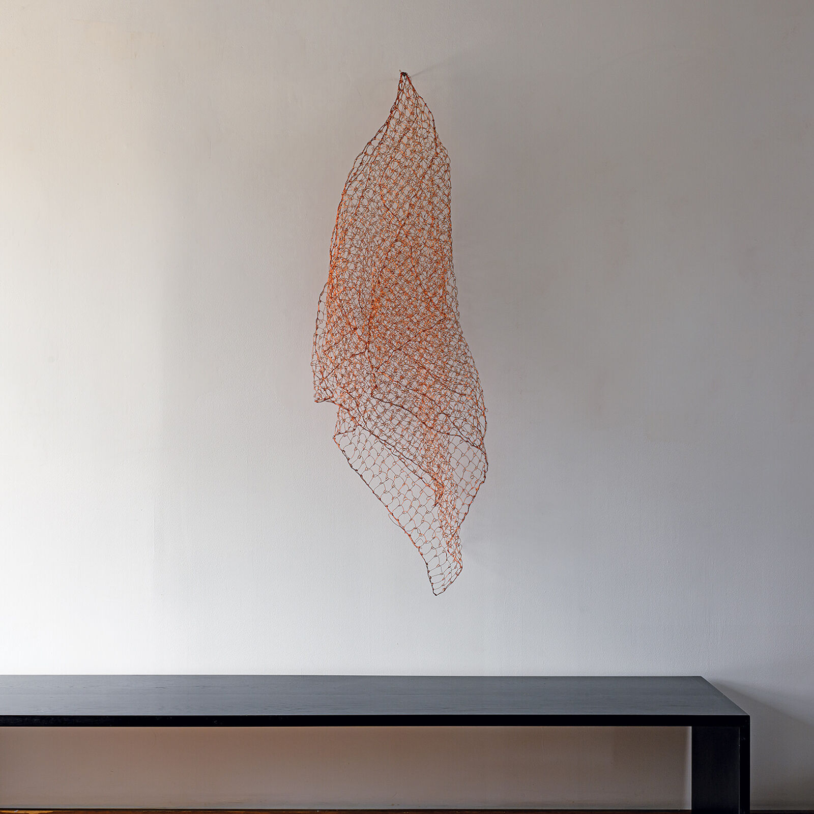 A minimalist interior scene featuring a suspended copper wire mesh sculpture against a plain wall.