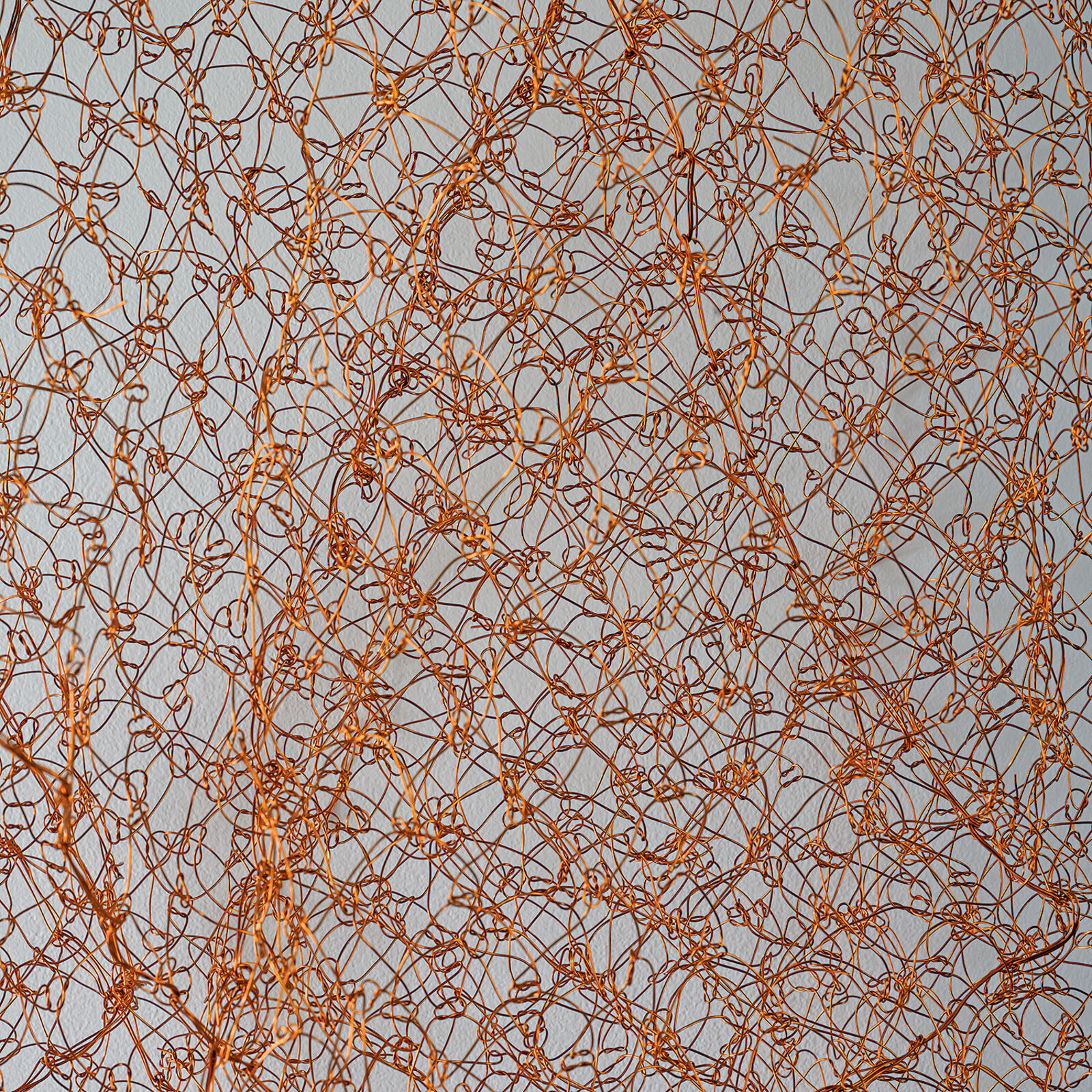 A minimalist interior scene featuring a suspended copper wire mesh sculpture against a plain wall.