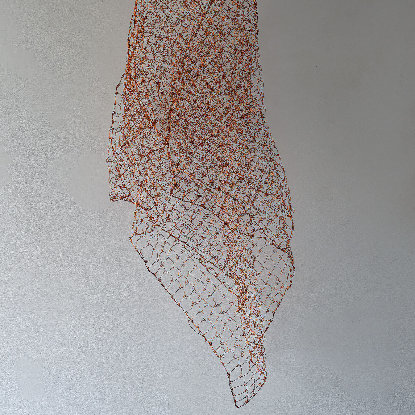 A minimalist interior scene featuring a suspended copper wire mesh sculpture against a plain wall.