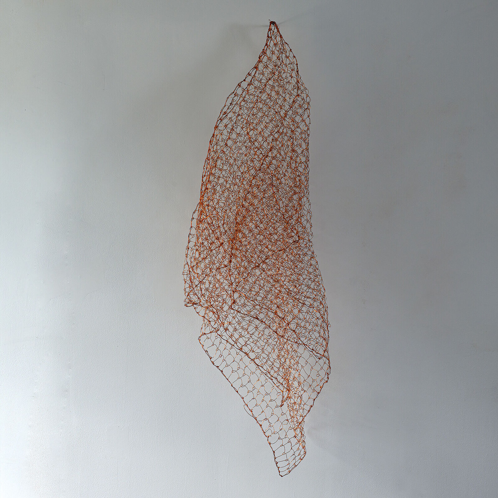 A minimalist interior scene featuring a suspended copper wire mesh sculpture against a plain wall.