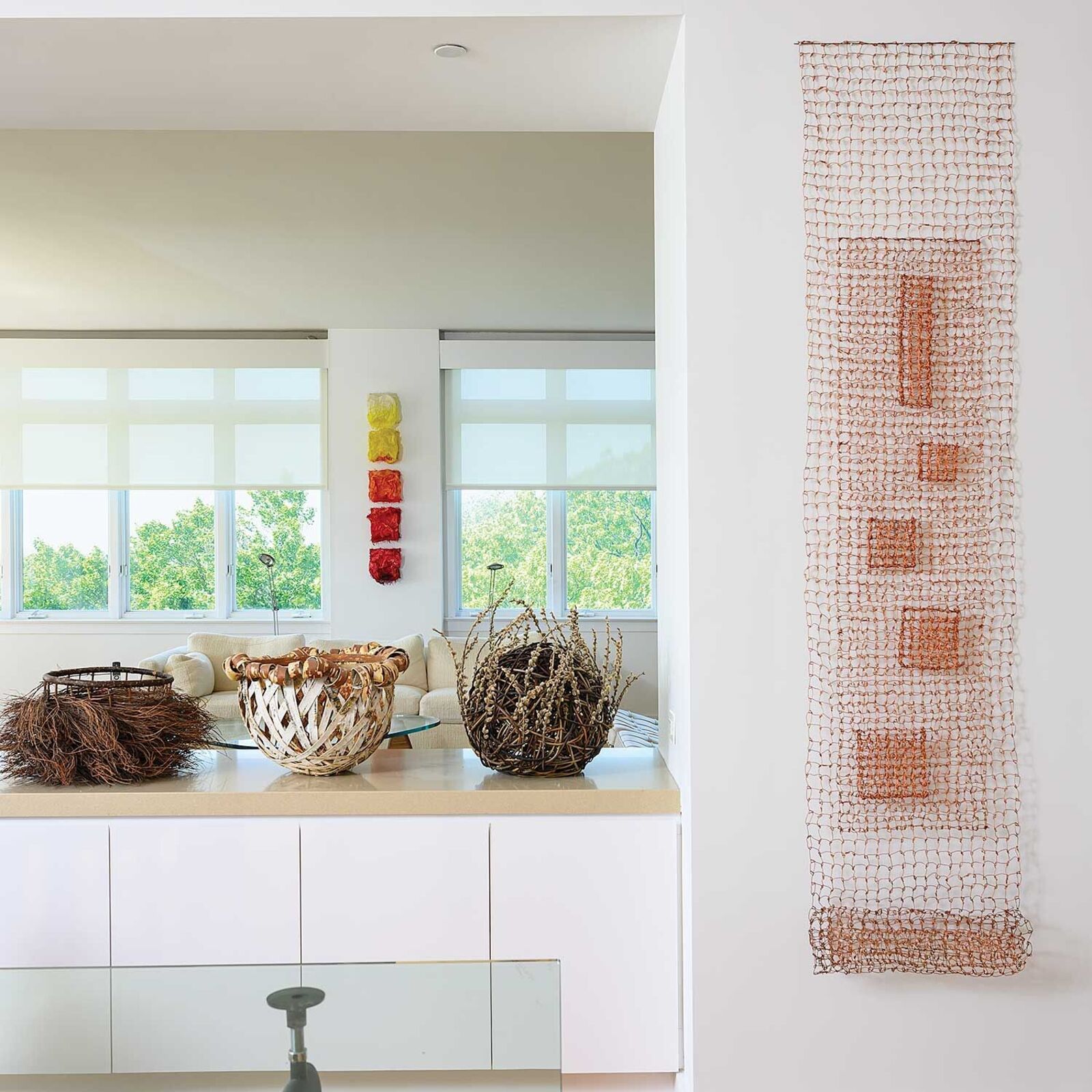 A minimalist wire mesh sculpture featuring a central vertical column of geometric shapes against a plain white wall.
