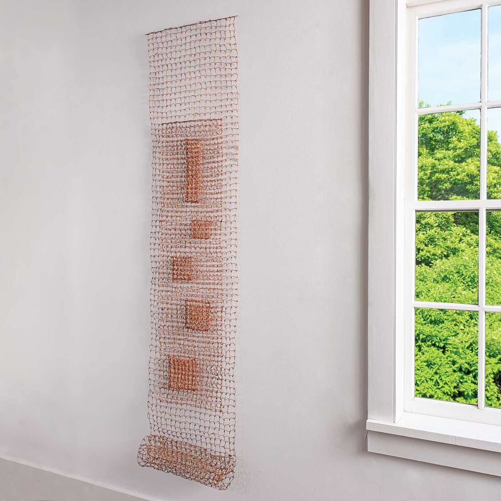 A minimalist wire mesh sculpture featuring a central vertical column of geometric shapes against a plain white wall.