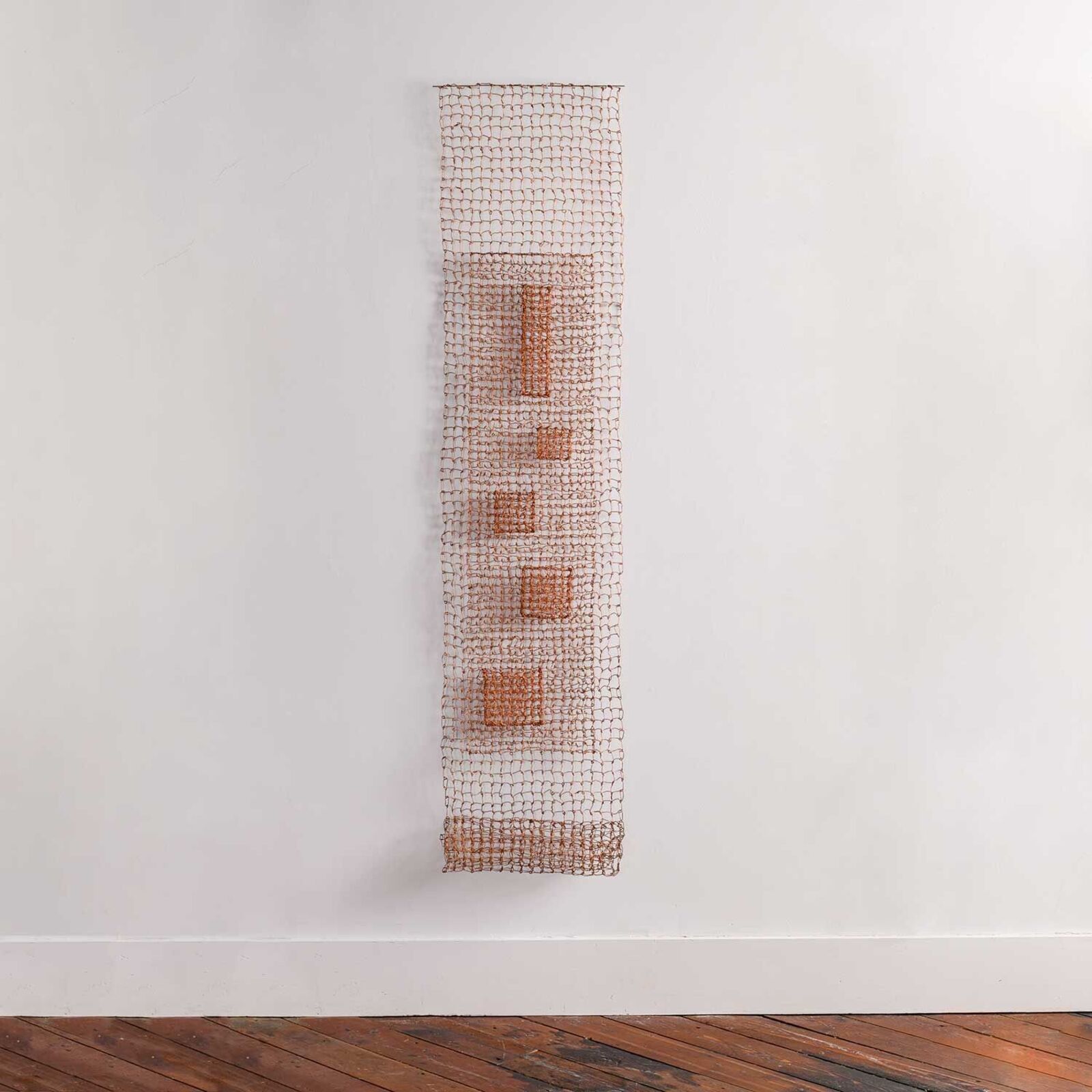 A minimalist wire mesh sculpture featuring a central vertical column of geometric shapes against a plain white wall.