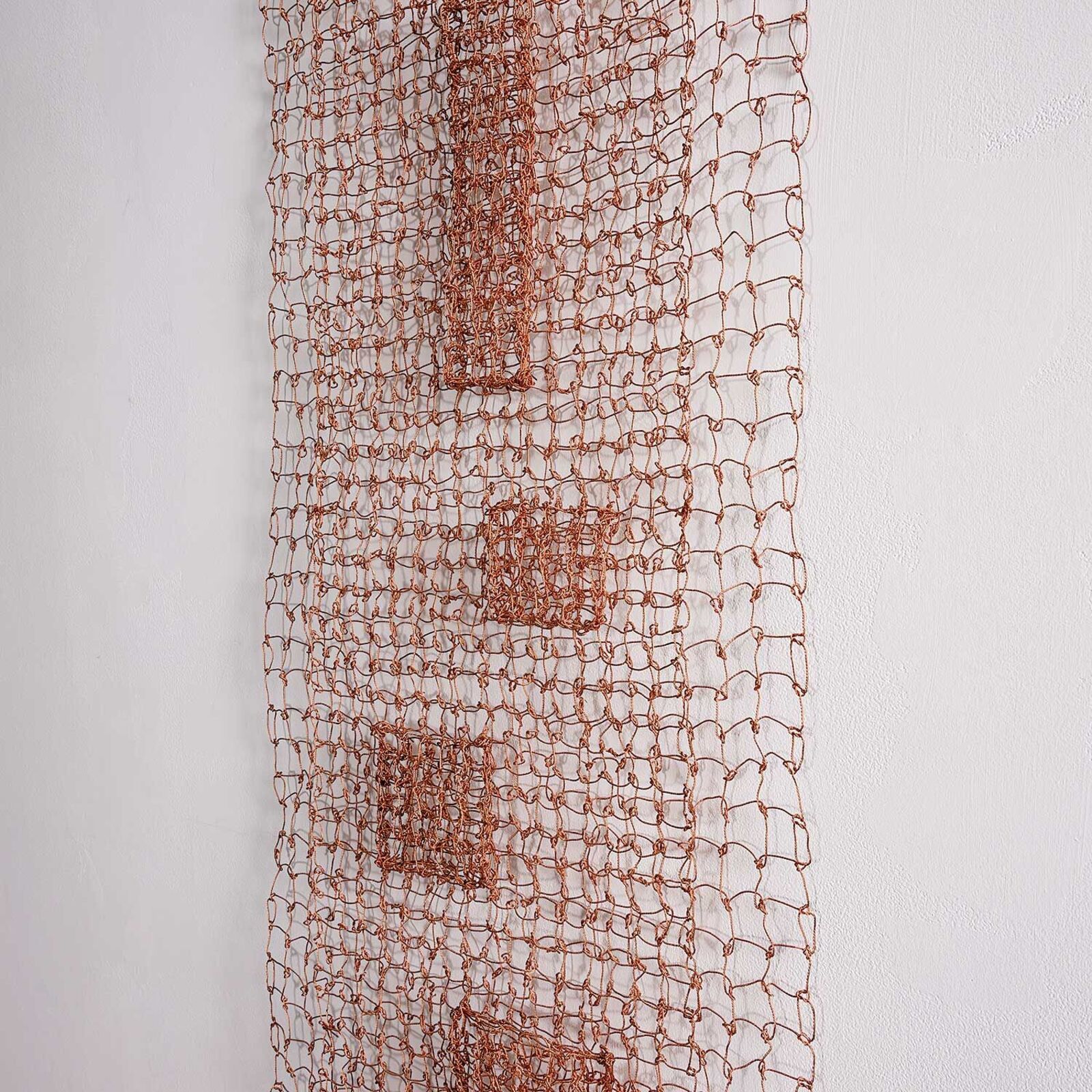 A minimalist wire mesh sculpture featuring a central vertical column of geometric shapes against a plain white wall.
