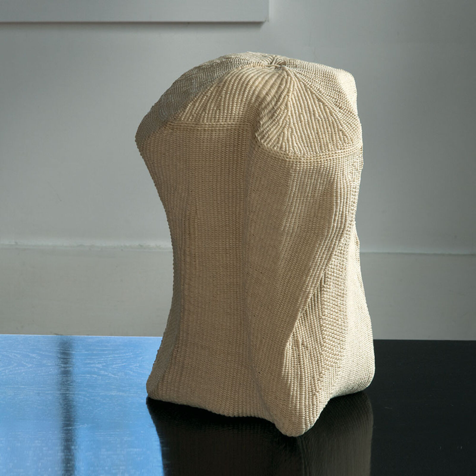 A sculptural beige chair cover with a ribbed texture, draped over an unseen form and resting on a glossy black surface.