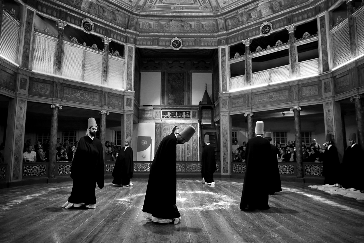A black-and-white photograph captures a group of robed figures performing a ritual dance inside an ornate, multi-level hall.