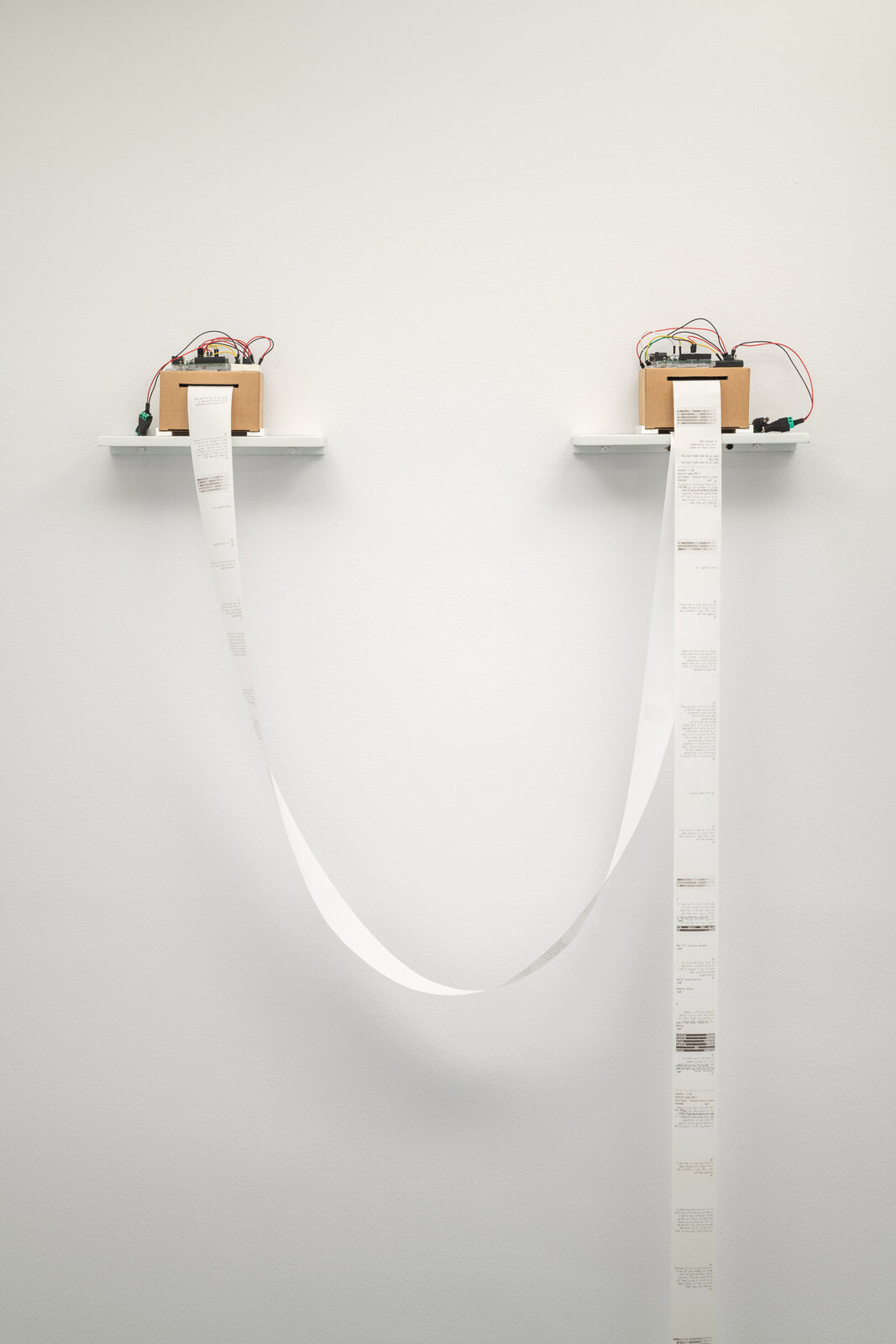 A minimalist installation featuring two wall-mounted devices with long paper strips hanging down.