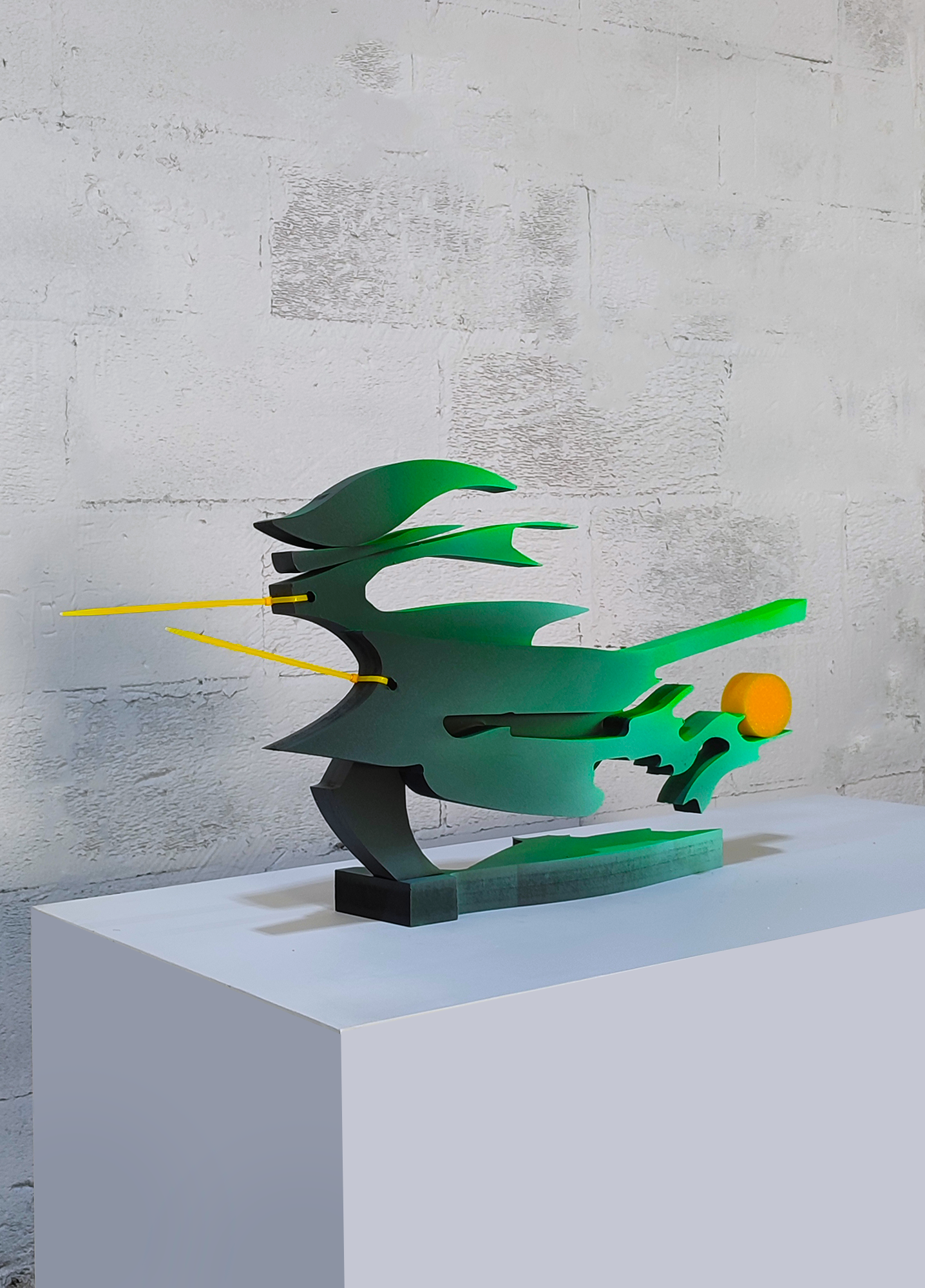 A green sculpture on a white pedestal in front of a white wall.