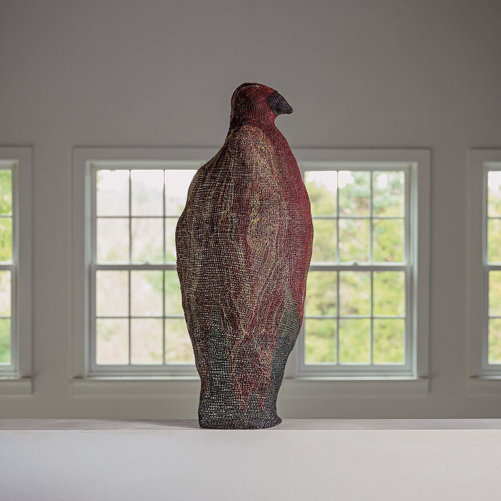 A textured, bird-shaped sculpture stands on a white surface in front of bright windows.