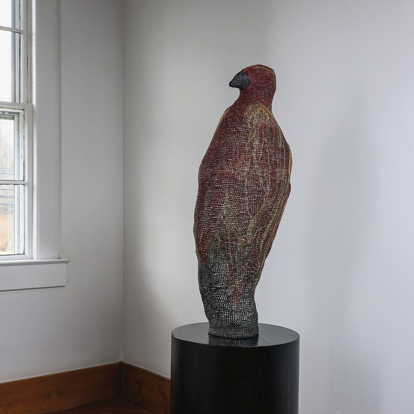 A textured, bird-shaped sculpture stands on a white surface in front of bright windows.