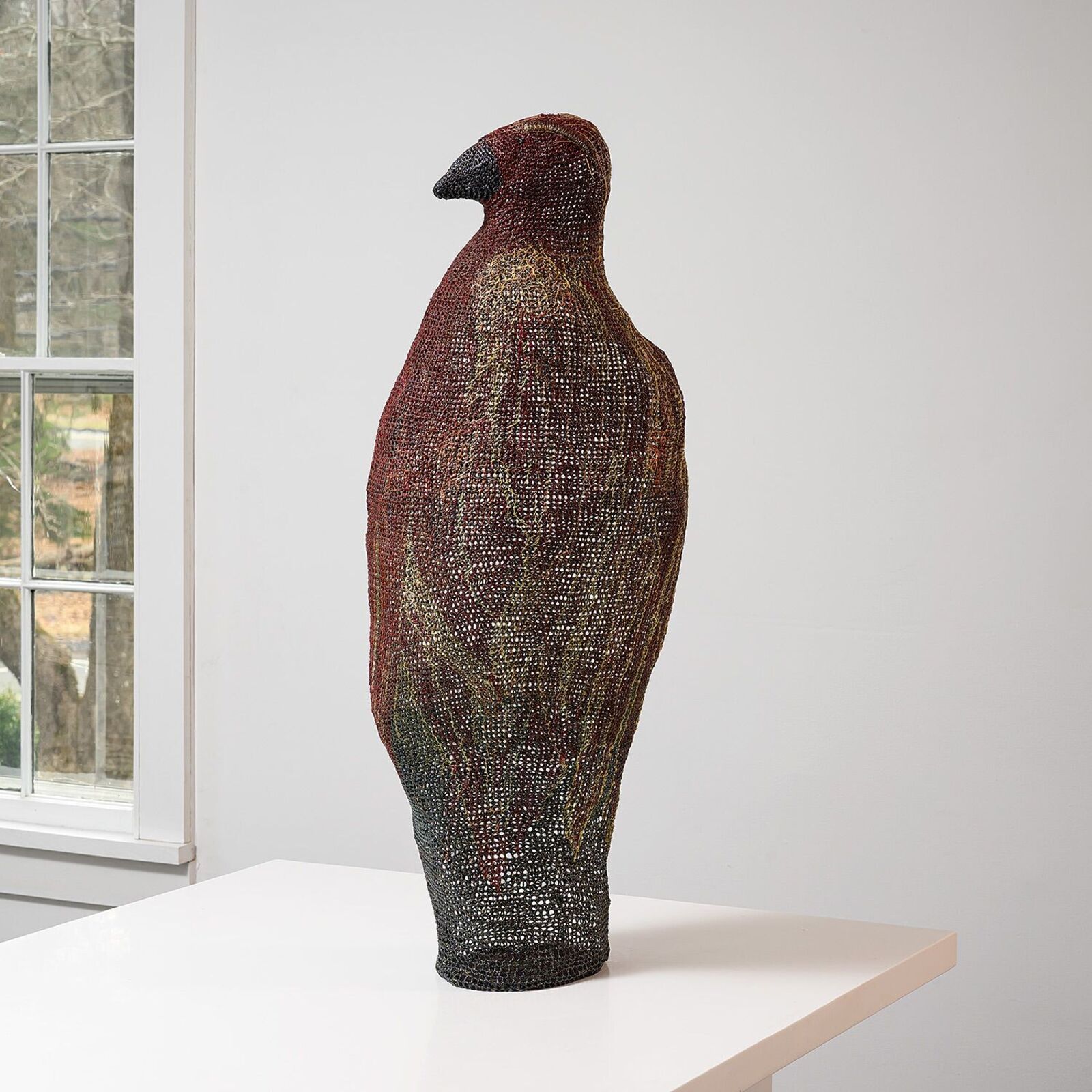 A textured, bird-shaped sculpture stands on a white surface in front of bright windows.