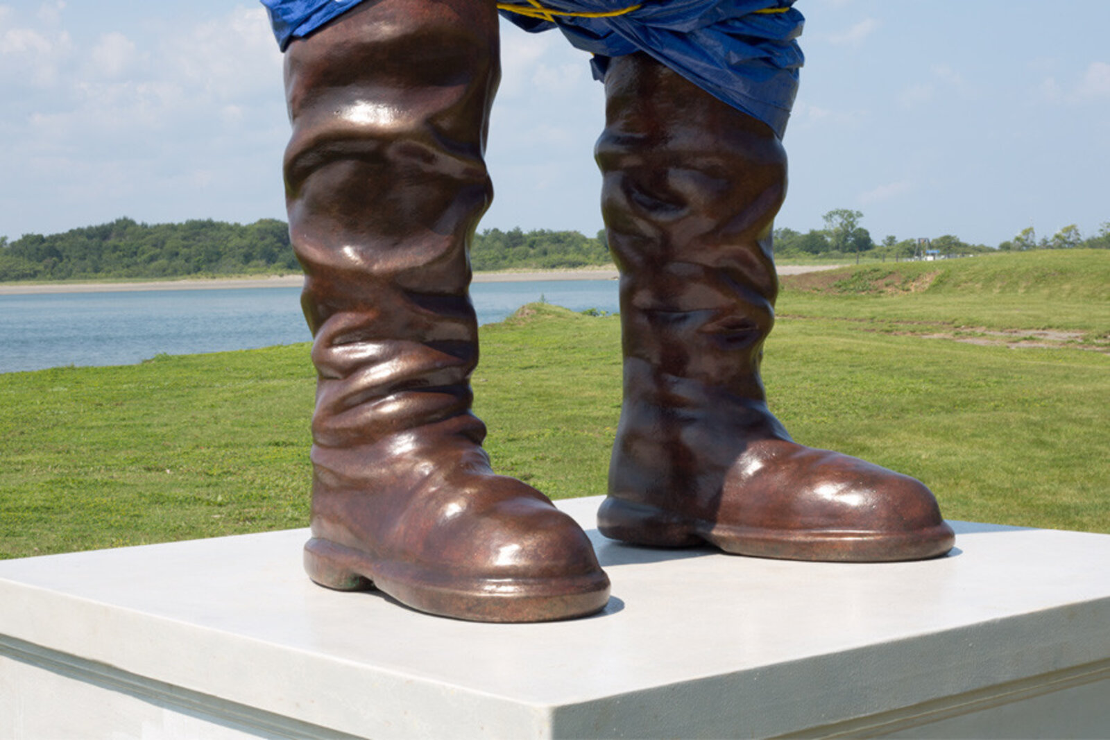 A surreal outdoor sculpture of a figure in blue plastic and boots standing on a pedestal.
