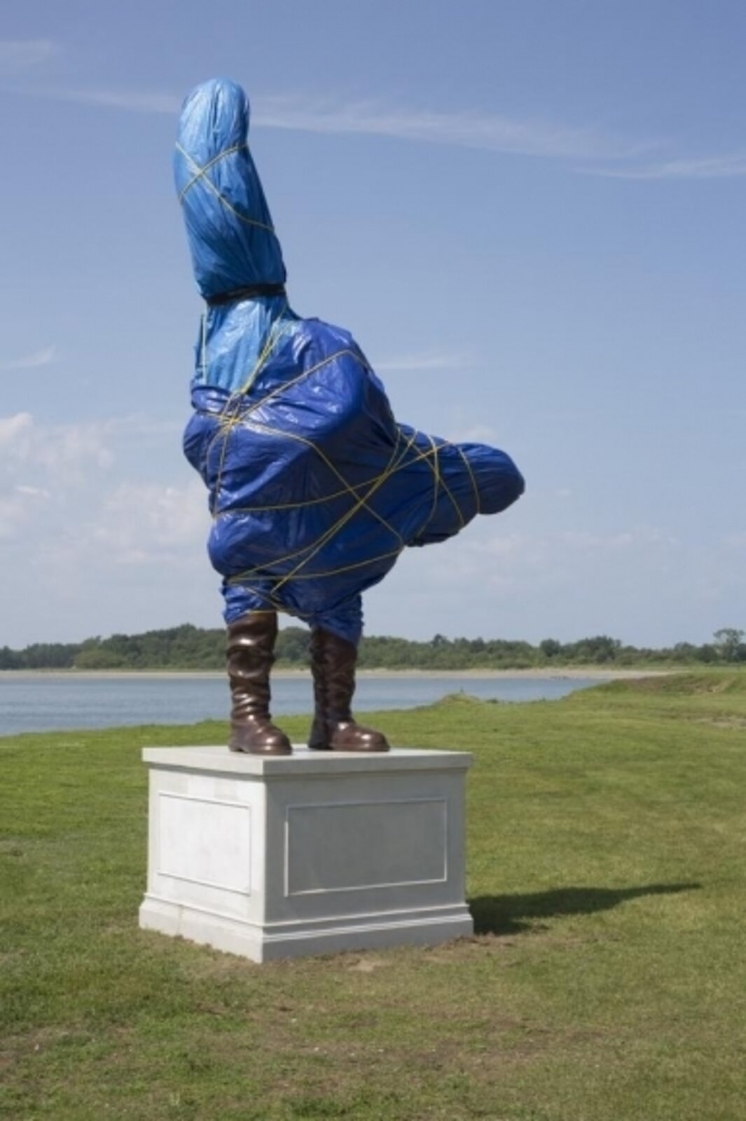 A surreal outdoor sculpture of a figure in blue plastic and boots standing on a pedestal.