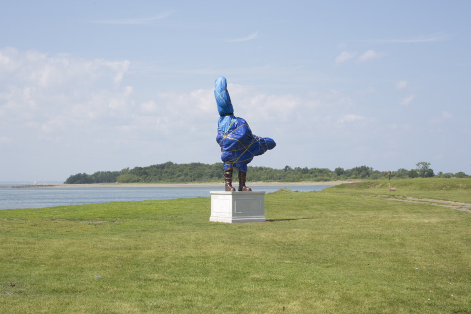 A surreal outdoor sculpture of a figure in blue plastic and boots standing on a pedestal.