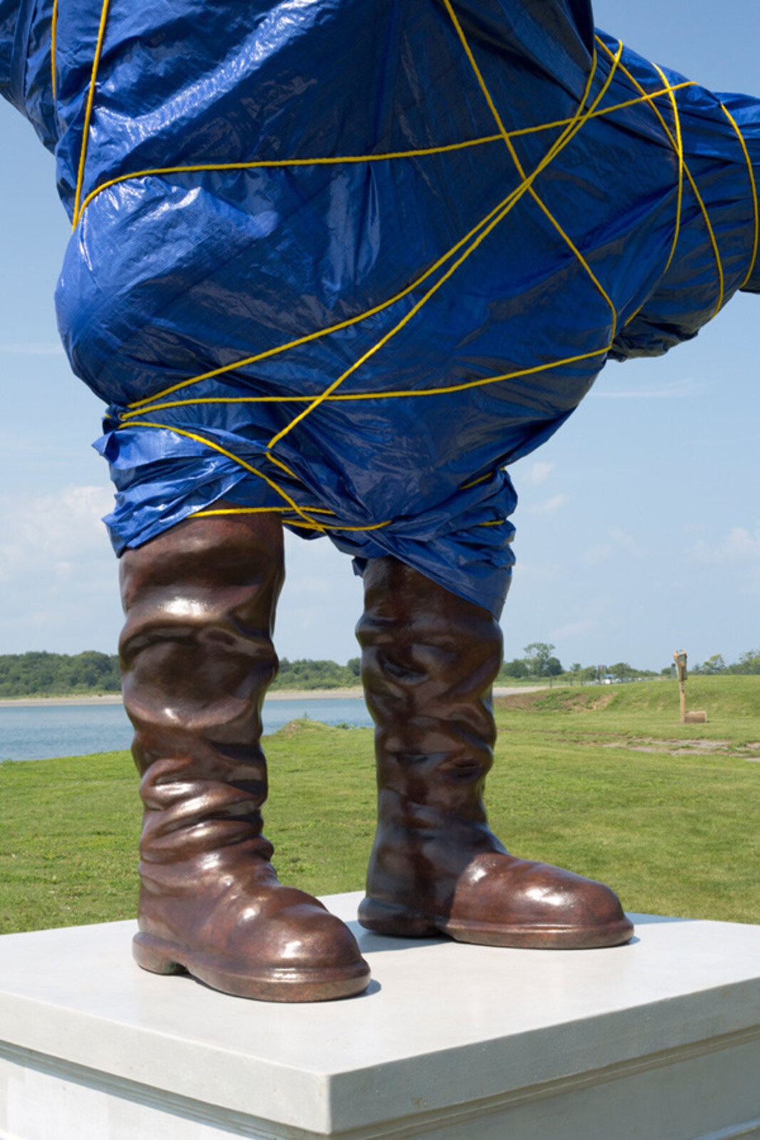A surreal outdoor sculpture of a figure in blue plastic and boots standing on a pedestal.