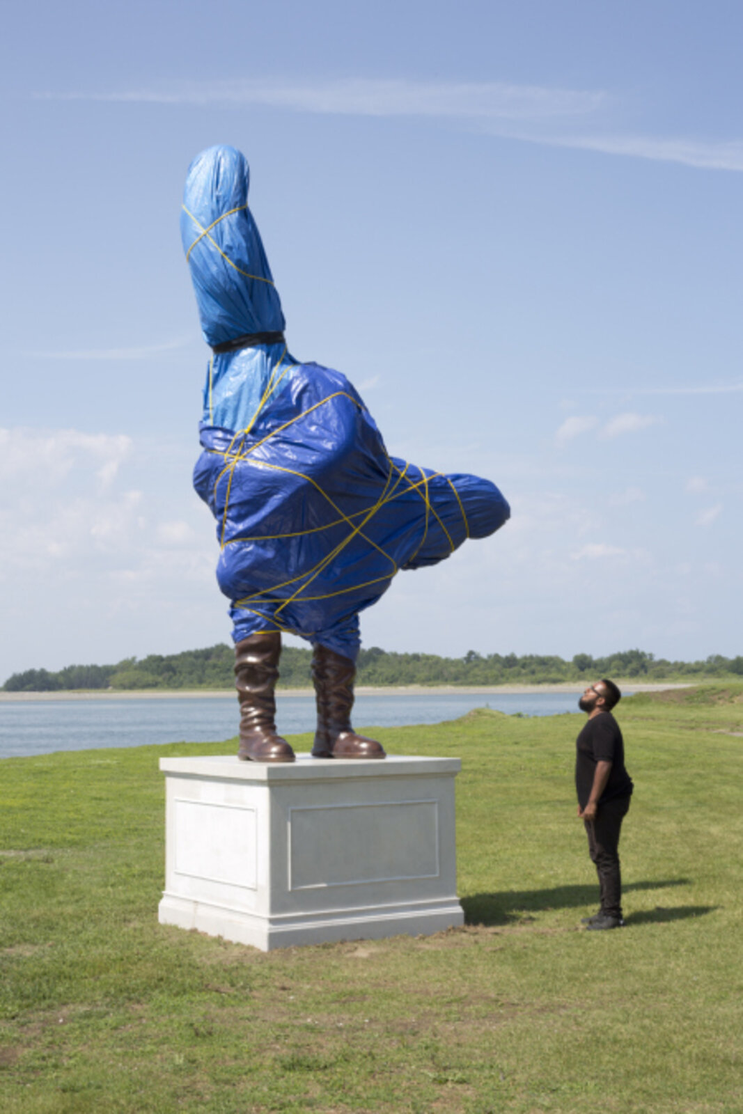 A surreal outdoor sculpture of a figure in blue plastic and boots standing on a pedestal.