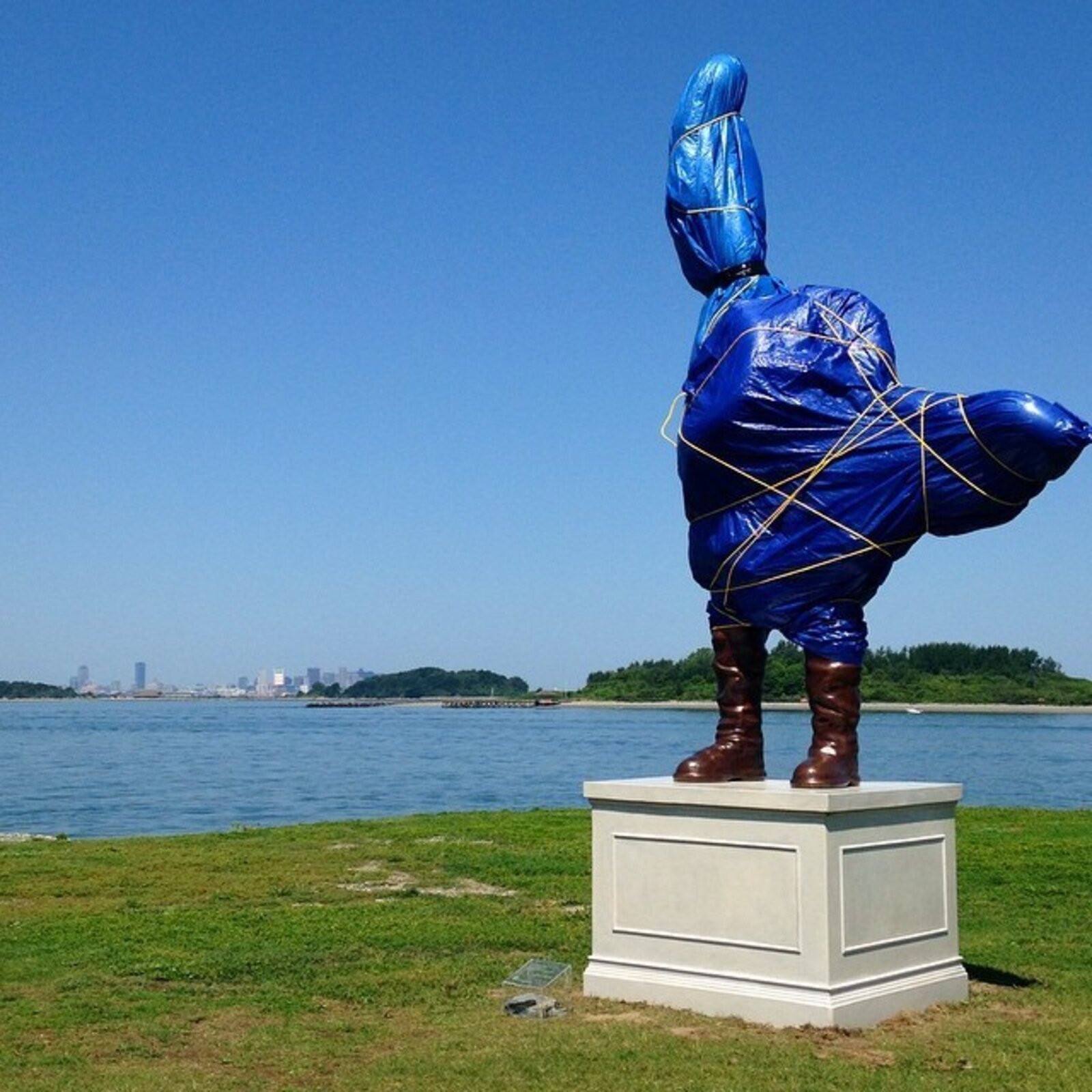 A surreal outdoor sculpture of a figure in blue plastic and boots standing on a pedestal.