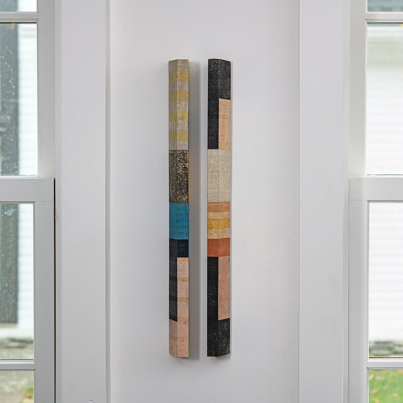 A vertical wall art piece featuring two elongated wooden slats with a patchwork of colorful, textured rectangular segments.