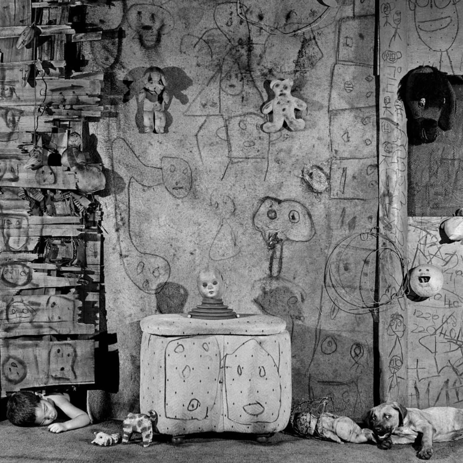 A black and white photograph of a cluttered room filled with toys, drawings, and a sleeping child.