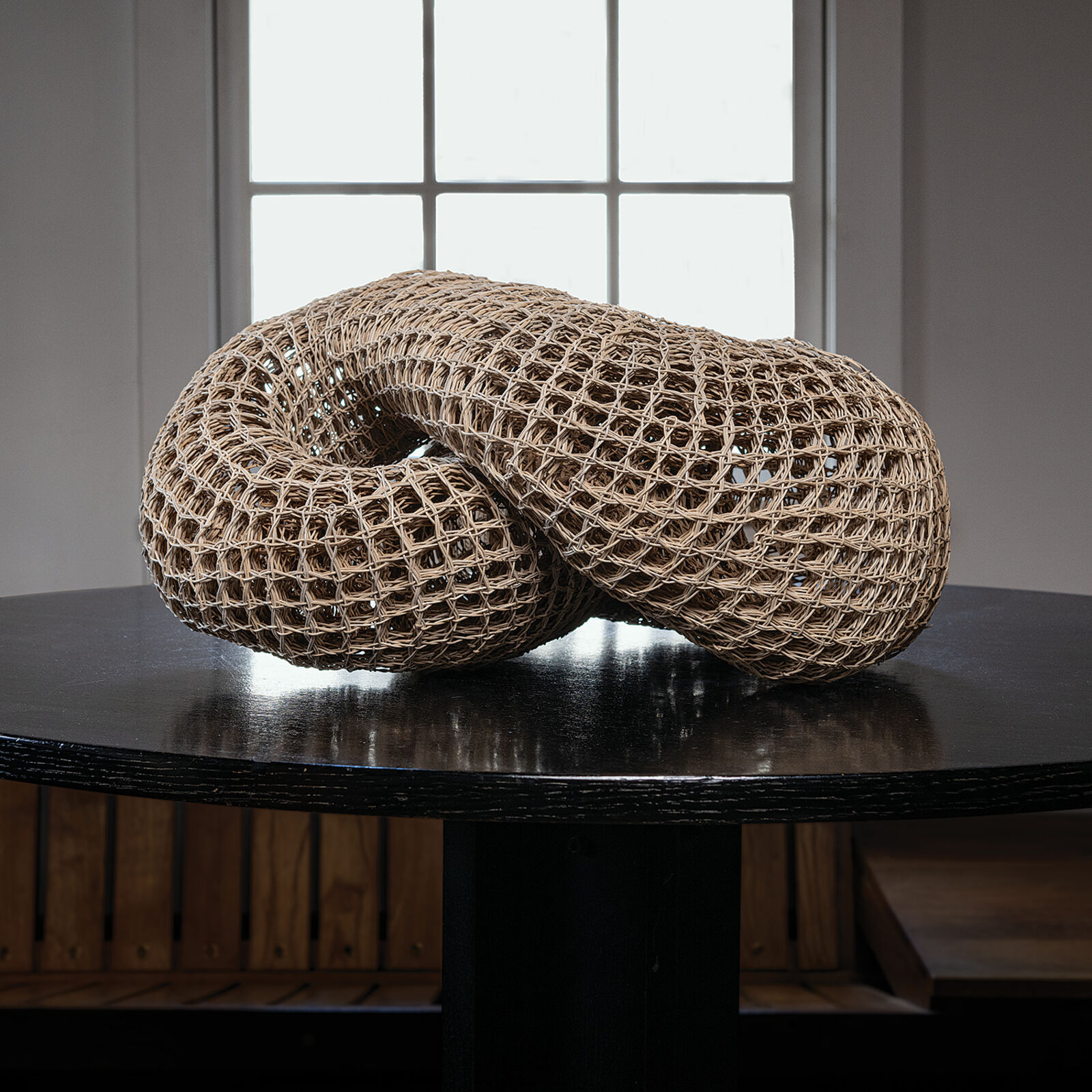 A woven, knot-shaped sculpture rests on a dark table in front of a bright window.