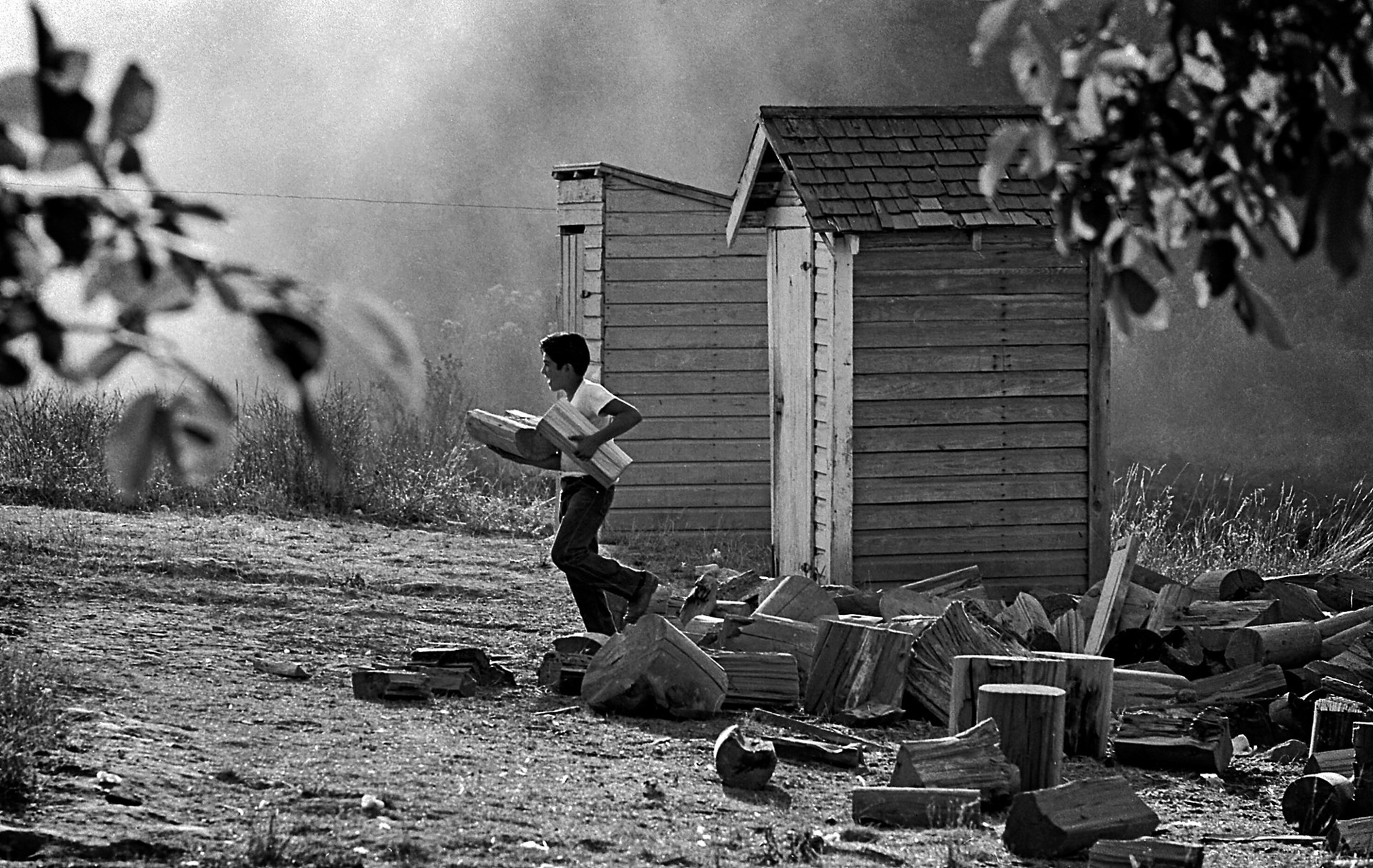 A man carrying a box of wood in front of a shack.