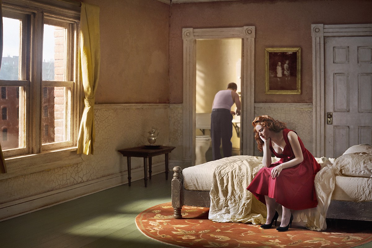 A woman in a red dress sitting on a bed in a room.