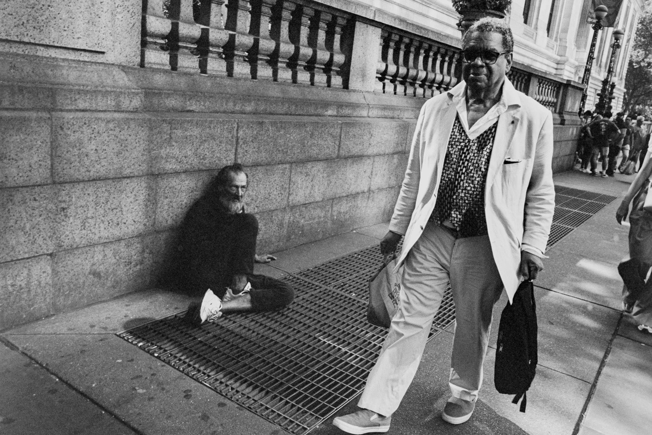 A man walking down a sidewalk next to a man sitting on the ground.