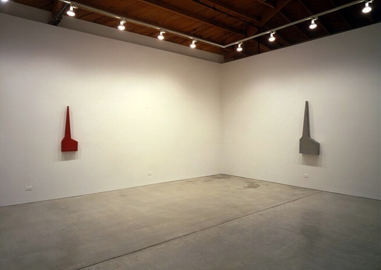 A minimalist sculpture featuring a sharp red spire rising from a flat base against a plain wall.